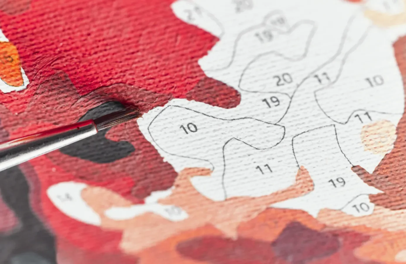 Close-up of a paintbrush pointing at a paint-by-numbers canvas with various sections numbered, featuring a red and black painted area, and unpainted areas with numbers outlined for painting.