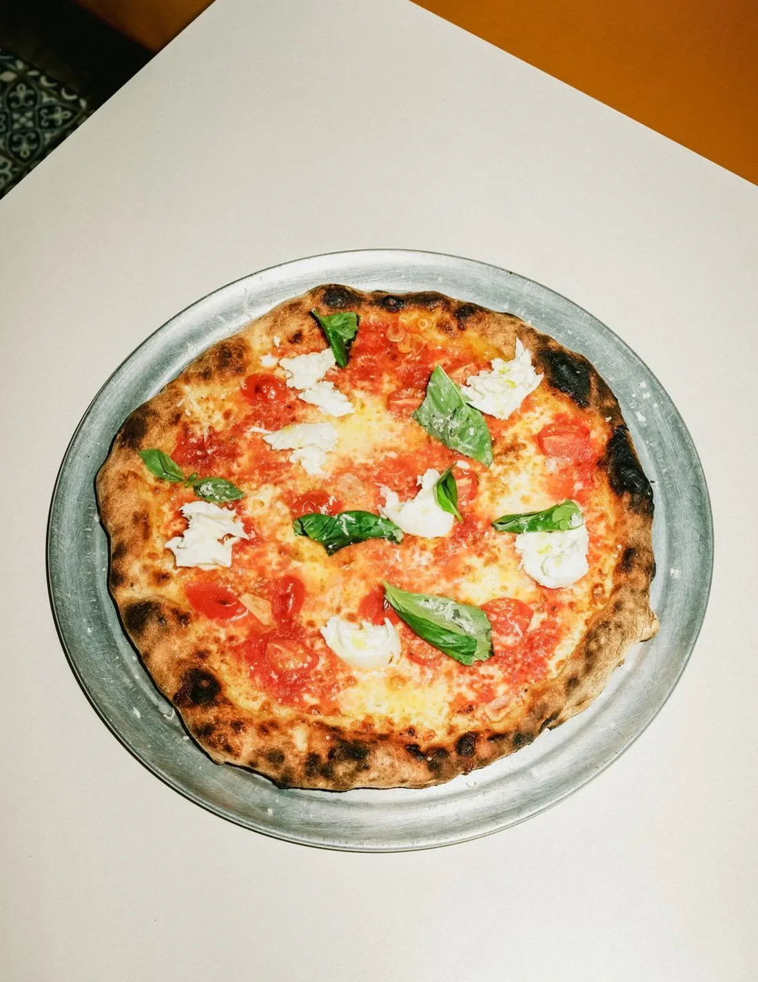 A pizza on a metal tray with a thin, slightly charred crust, topped with tomato sauce, mozzarella cheese, cherry tomato slices, and fresh basil leaves.