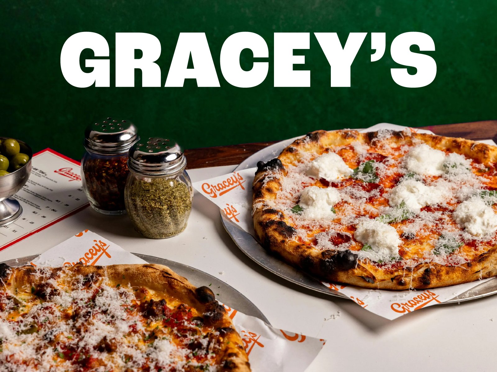 Two pizzas on a table, one with cheese and a second partially visible with toppings, with salt and pepper shakers, and a small bowl of green olives, in front of a green wall with the words 'Gracey's' in large white letters.