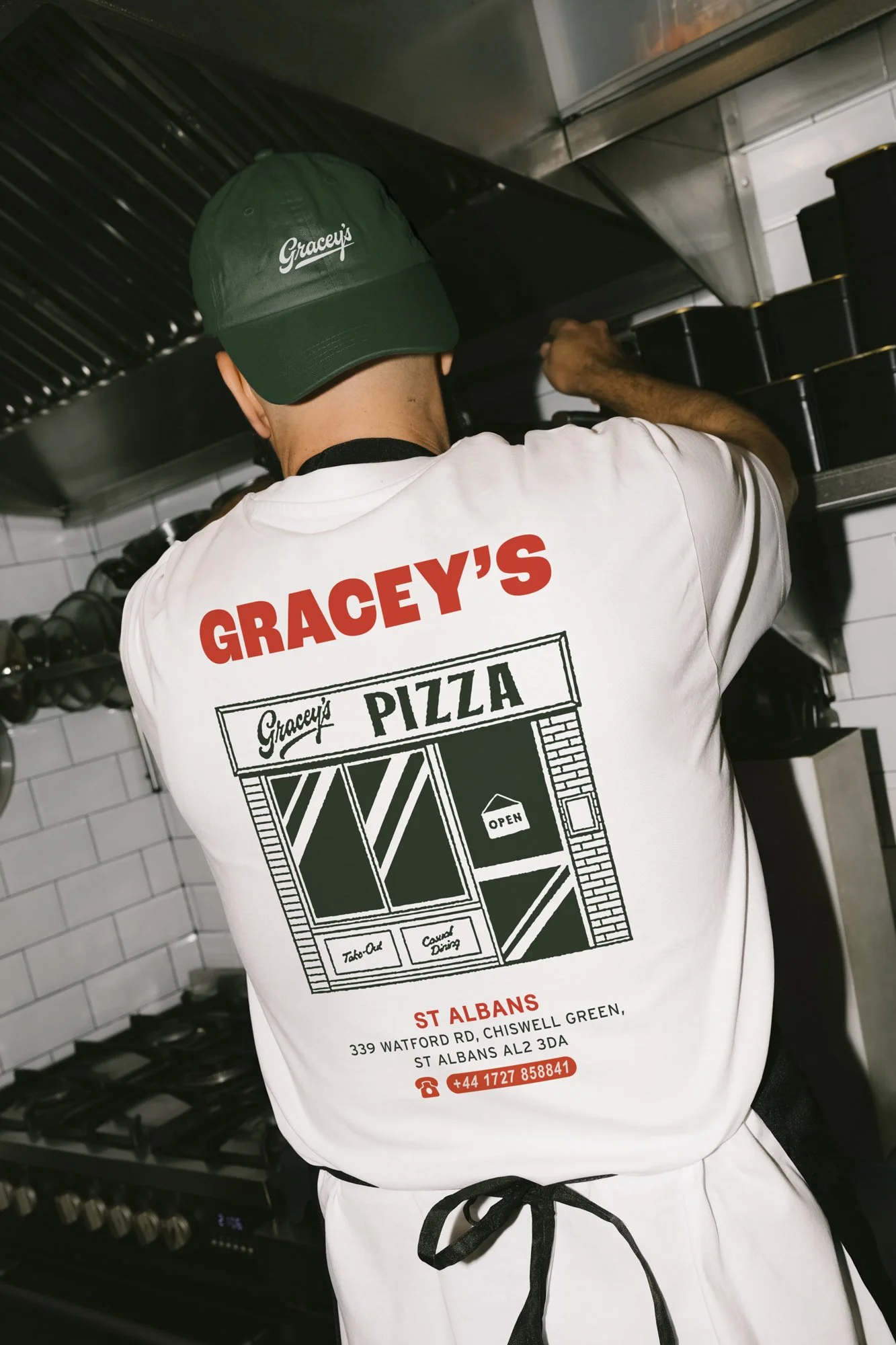 Person wearing a white Grimacy's Pizza t-shirt and a green cap, working behind a kitchen grill. The t-shirt has a graphic image of a pizza shop storefront and address details.