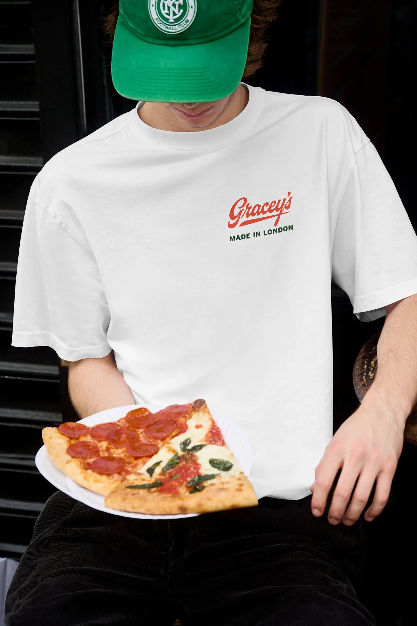 A person wearing a green cap with a football club logo and a white T-shirt with 'Gracey's Made in London' written on it is holding a plate with two slices of pizza. One slice has pepperoni and the other has tomato, basil, and cheese.