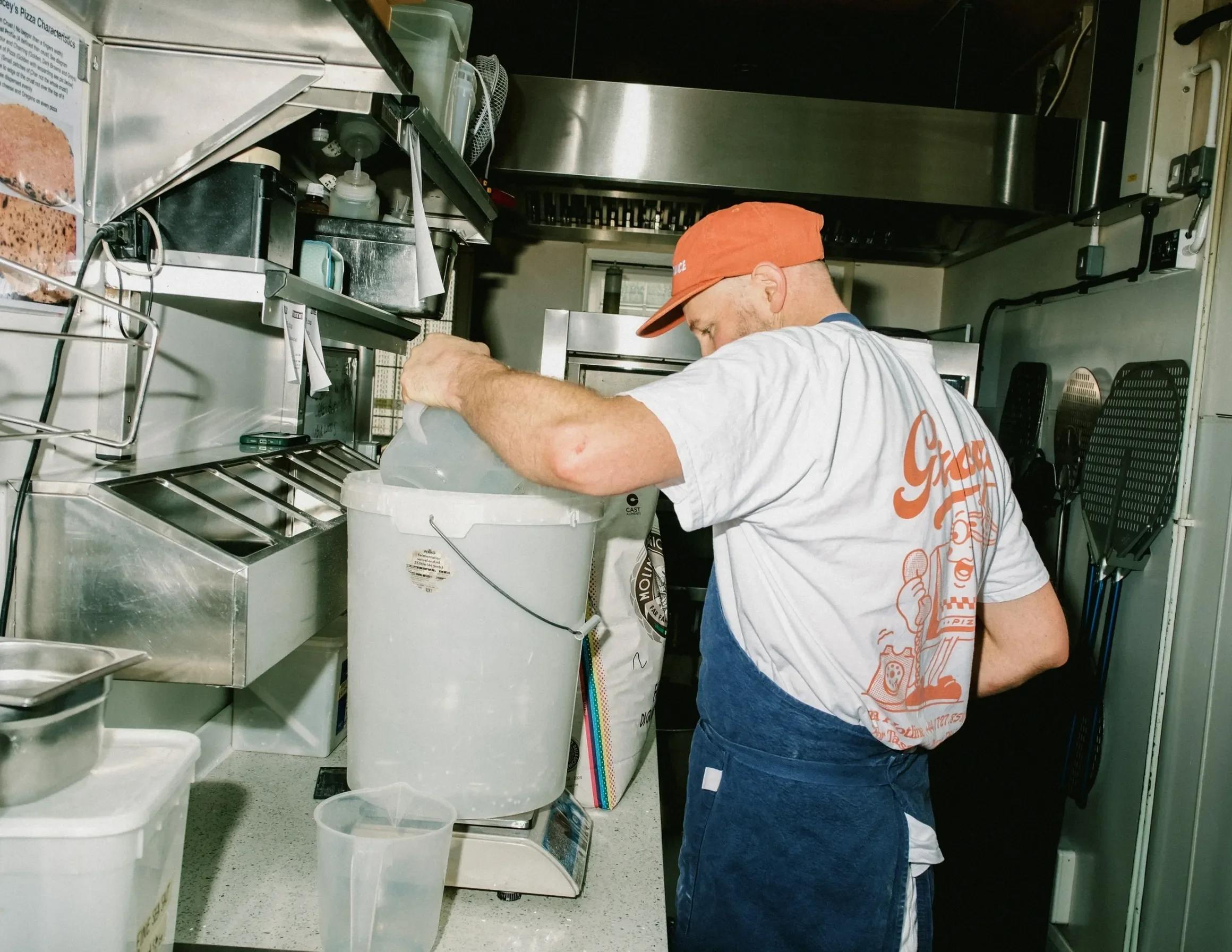 A person wearing a red cap and gray T-shirt working in a commercial kitchen, surrounded by kitchen equipment, containers, and utensils.