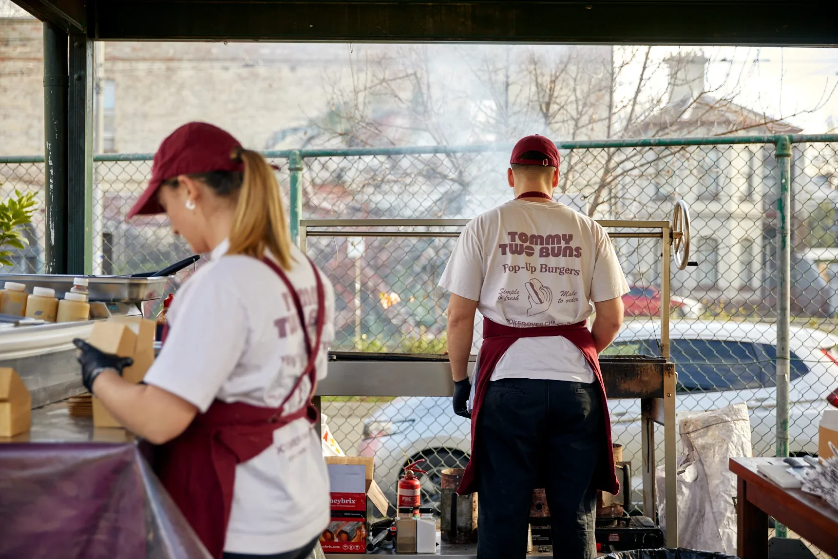 Two Tommy Buns workers cooking burgers over a grill, wearing their designed merch.
