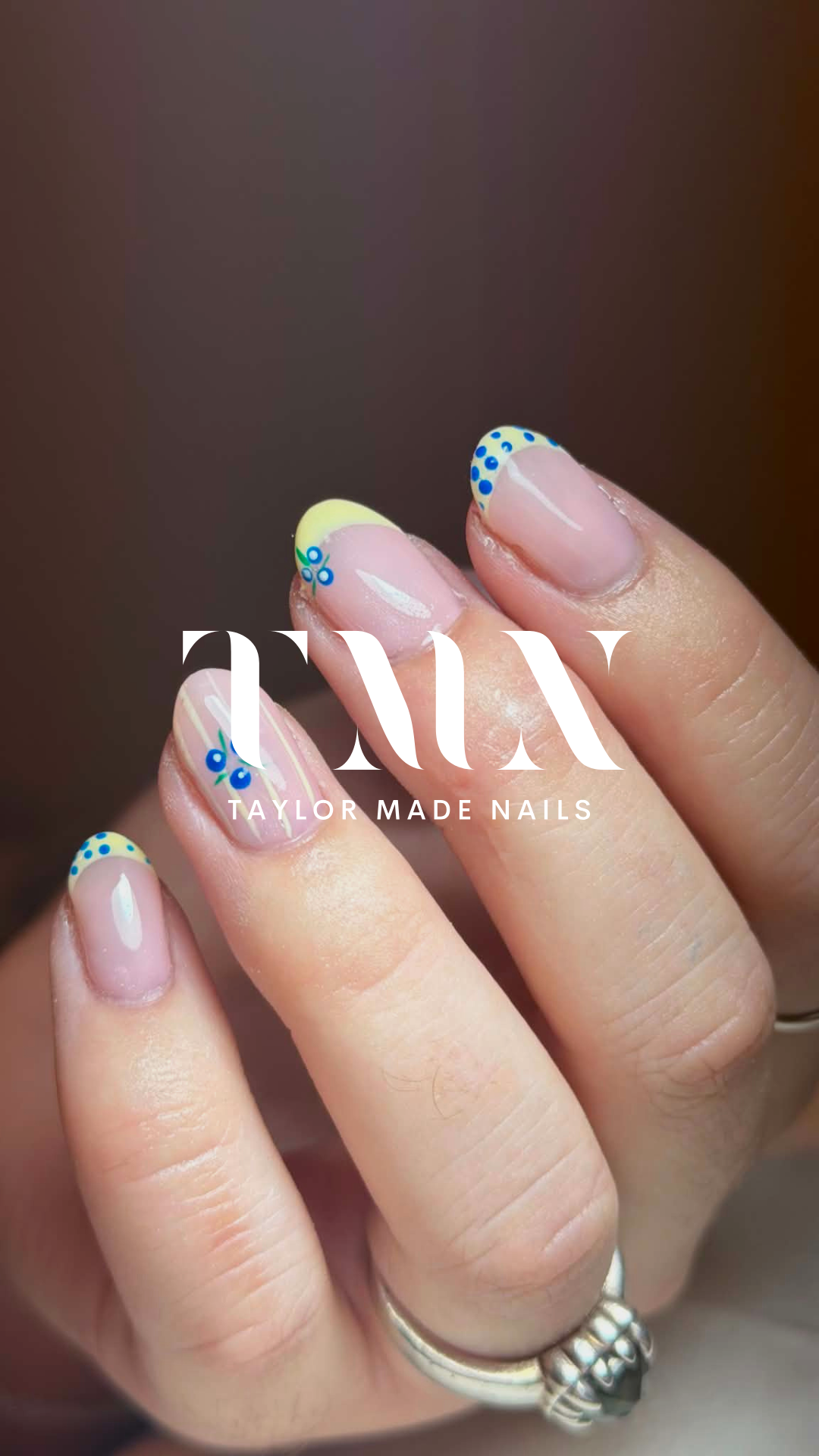 Close-up of a hand with manicured nails featuring yellow tips and blue dots, and pink nails with white lines, with a white logo reading 'TAYLOR MADE NAILS' overlayed on the image.
