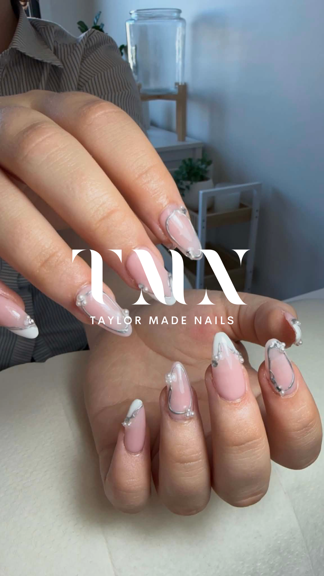 Close-up of a person's hands with decorated acrylic nails, featuring a clear base with white and silver accents and small pearl-like embellishments. In the background, there is a person wearing a striped shirt, and a blurred interior setting with a g