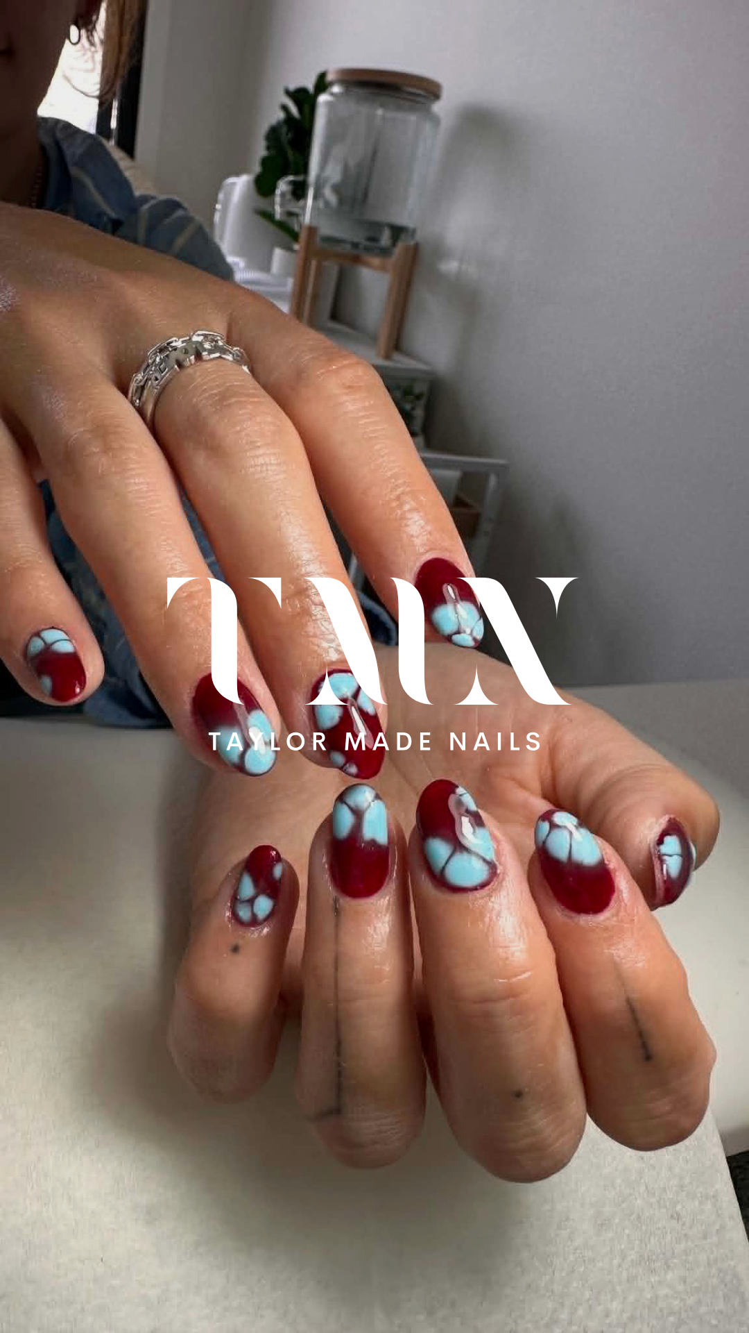 Close-up of a person's hands showing freshly painted nails with a red and blue swirl design, wearing a silver ring. In the background, a person with dark hair, a plant, and a water dispenser are visible. The image has an overlay with the text 'TAYLOR