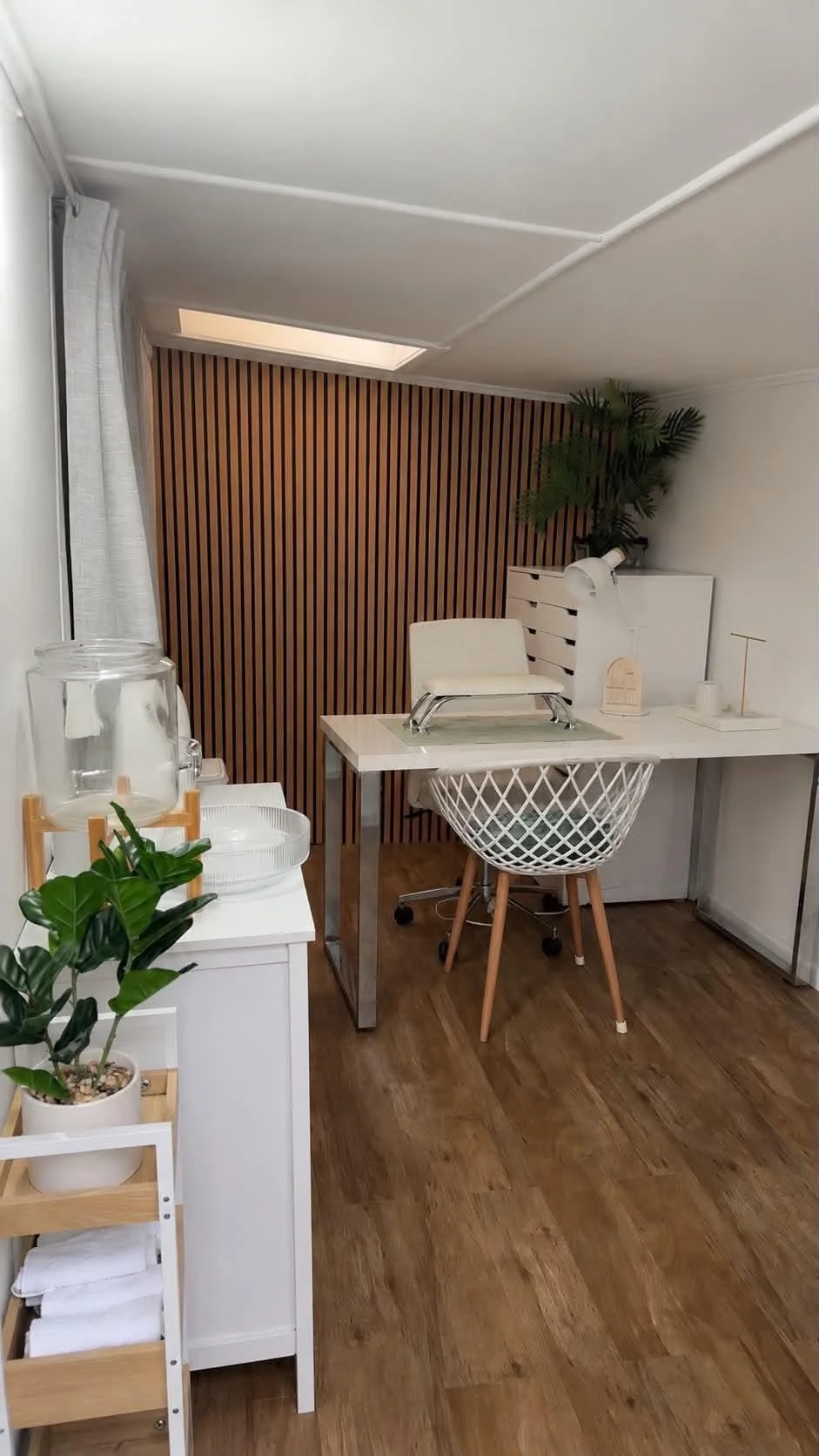 Interior of a modern, minimalist office or workspace with a white desk, ergonomic chair, wooden flooring, and a light wood slat wall accented by a large leafy plant and a skylight