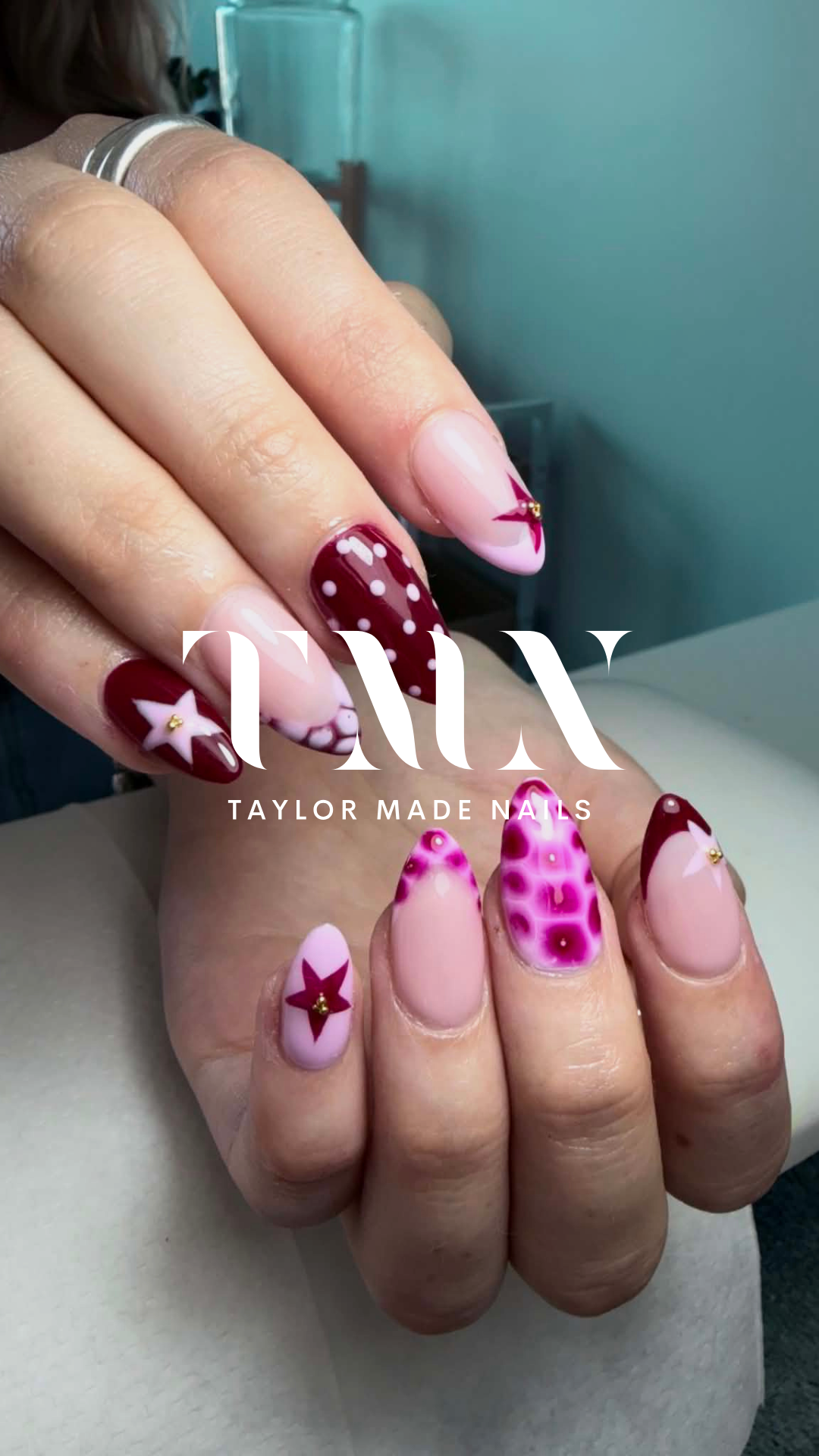 Close-up of hands with decorated nails featuring red, pink, and white holiday-themed designs, including stars, polka dots, and abstract patterns.