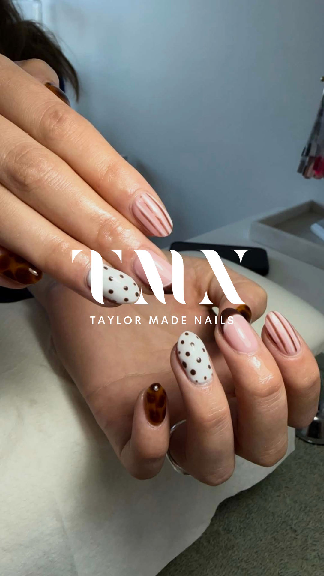 Close-up of hands with manicured nails featuring pink, white with dark dots, and brown tortoiseshell designs, with the logo 'TAYLOR MADE NAILS' overlayed.