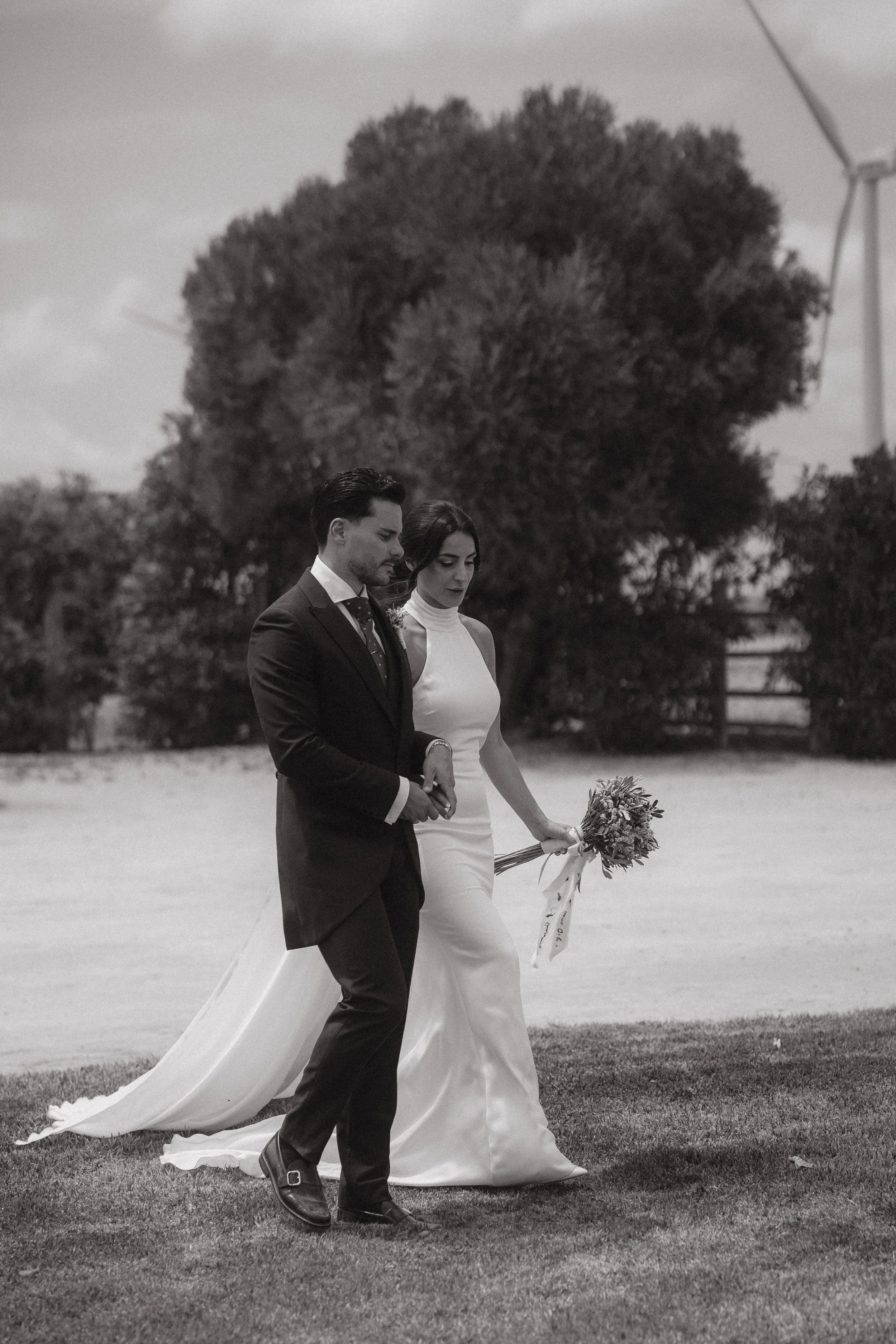 Pareja de recién casados caminando en un campo, la novia lleva vestido blanco y sostiene un ramo de flores, el novio viste traje negro, en un día nublado.