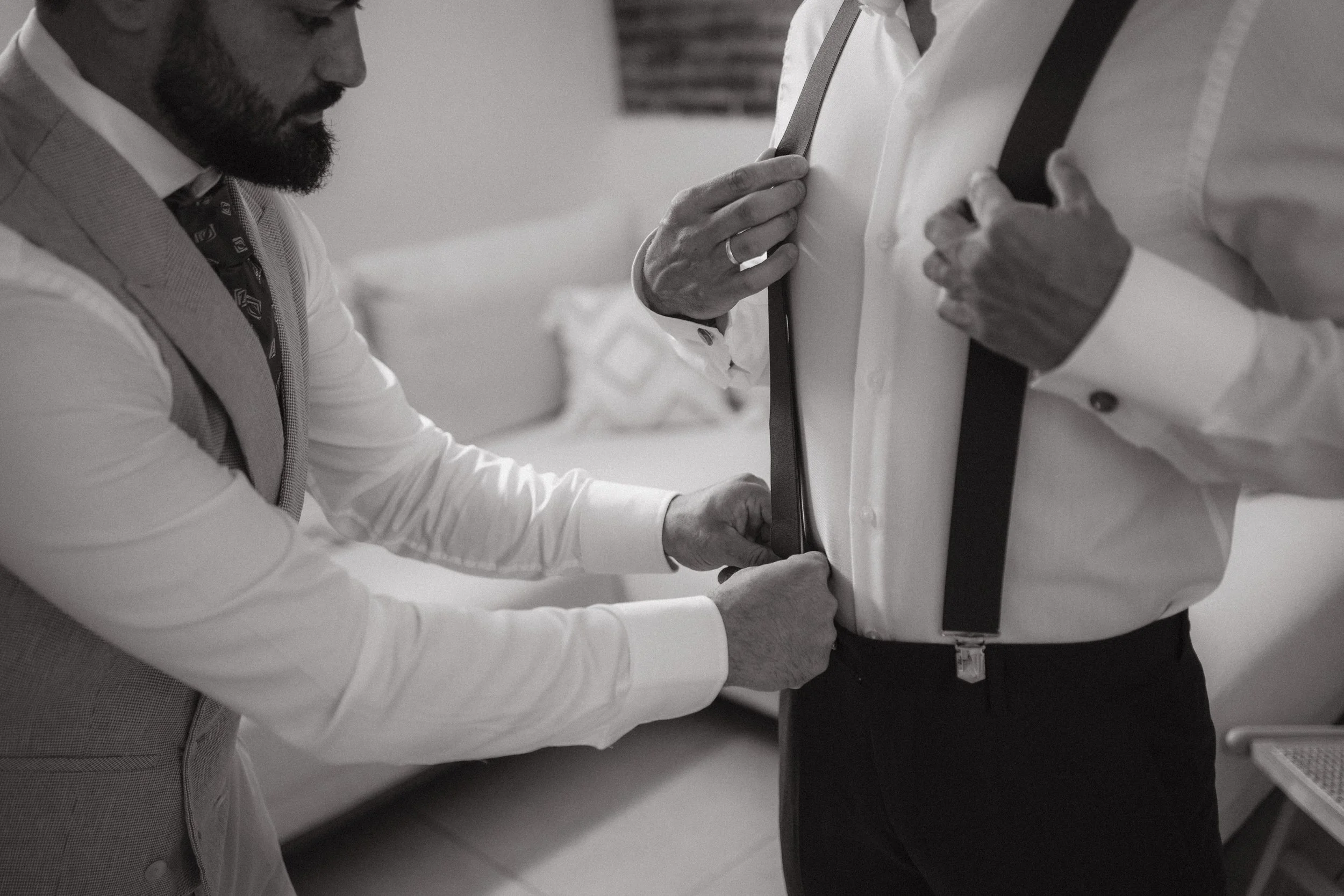 Dos hombres visten ropa formal, uno ajustándose una corbata y el otro ayudándole a ajustar una cinturilla o prenda en la cintura, en un entorno interior.