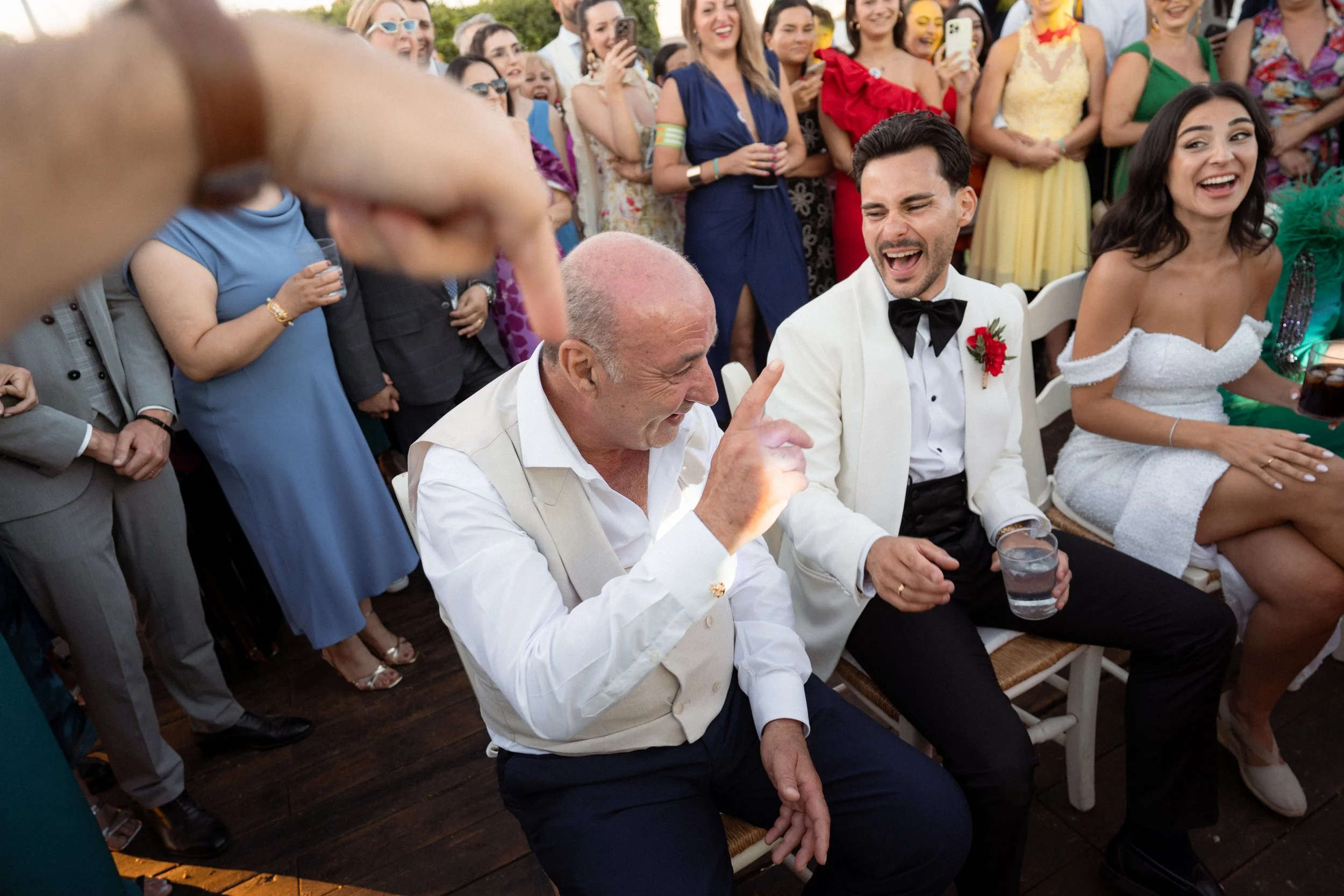 Grupo de personas en una celebración, con un hombre mayor y un hombre vestido de esmoquin en primer plano, riendo, y varias personas en el fondo, algunas tomando fotos.