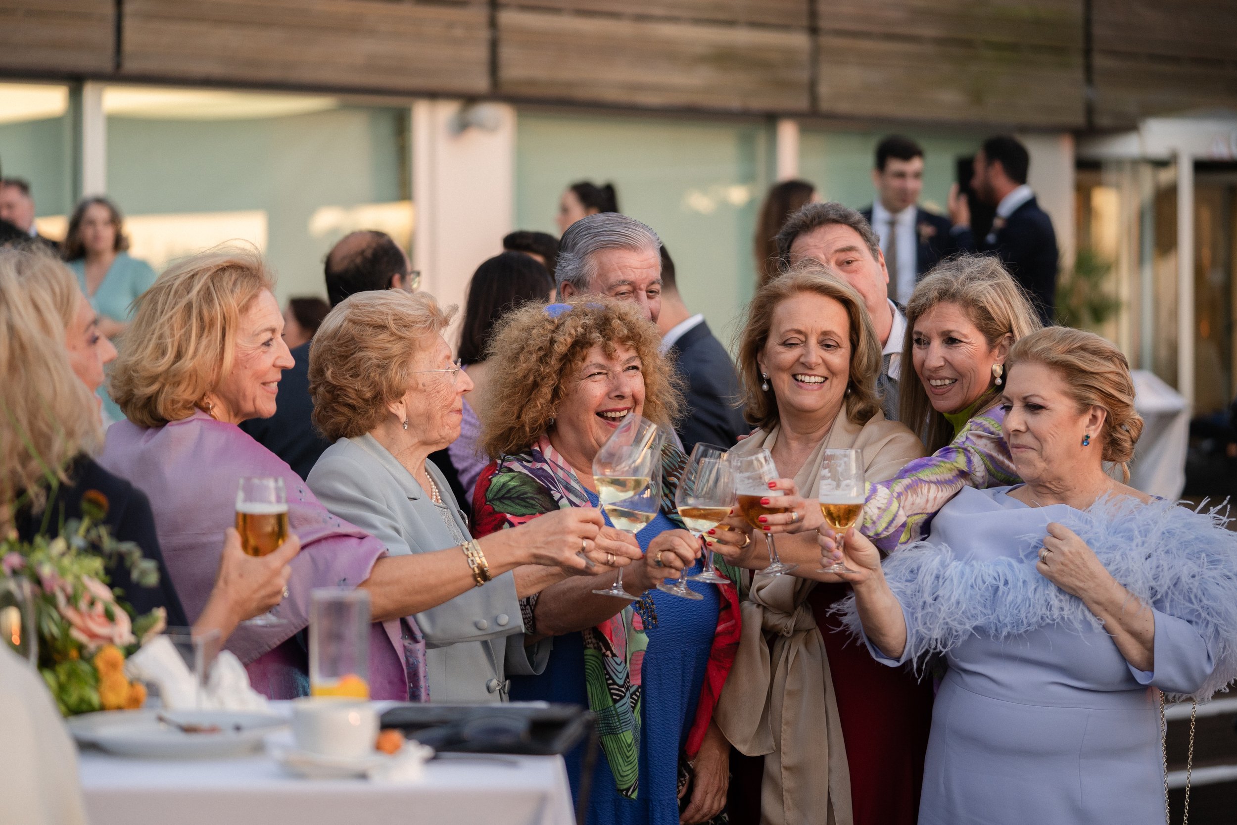 Grupo de personas mayores, principalmente mujeres, brindando con copas de vino junto a una mesa en un evento social al aire libre.