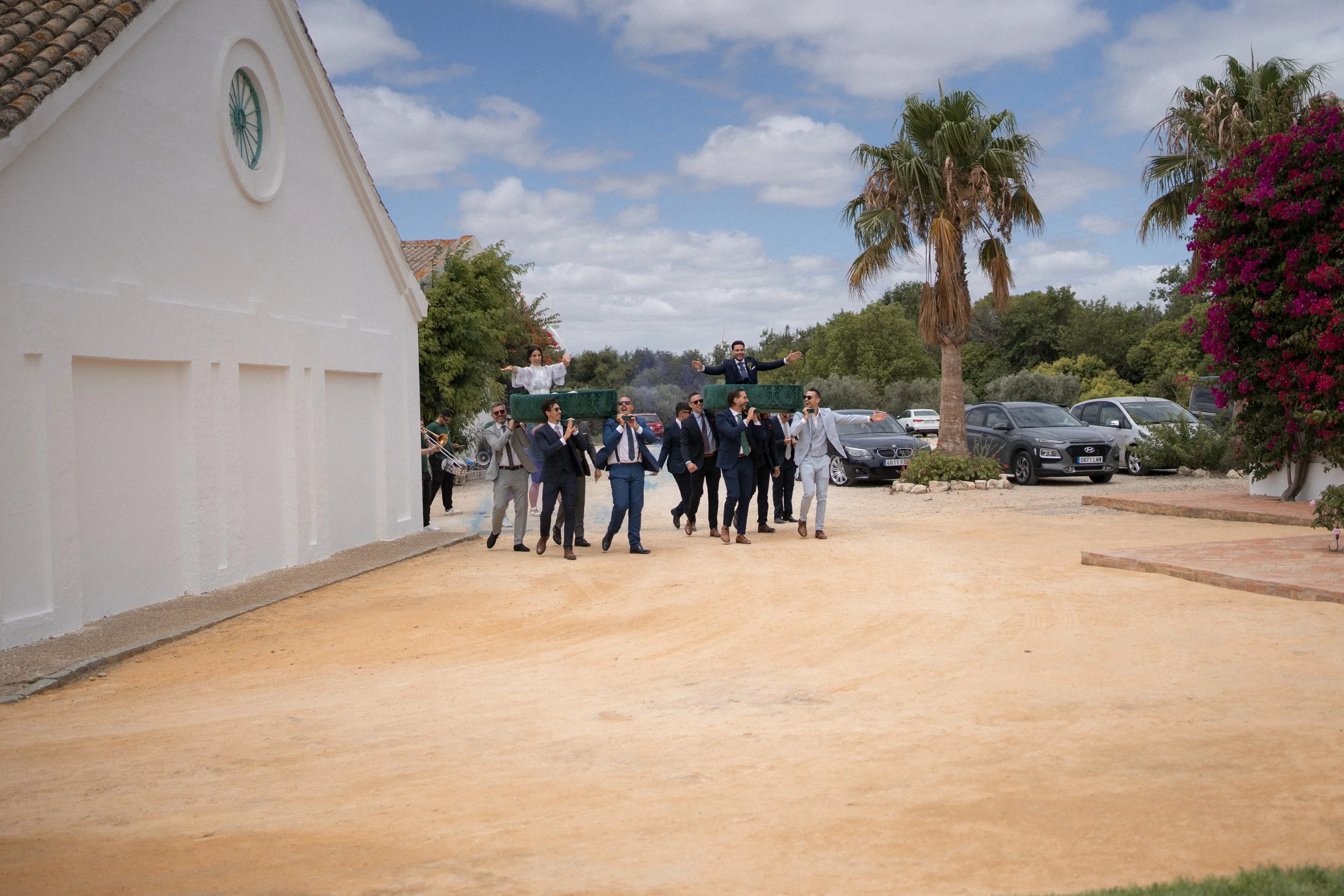 Un grupo de personas vestido formalmente participando en una procesión con un paso nupcial, mientras una banda toca música en un espacio abierto con árboles y carros estacionados en el fondo.