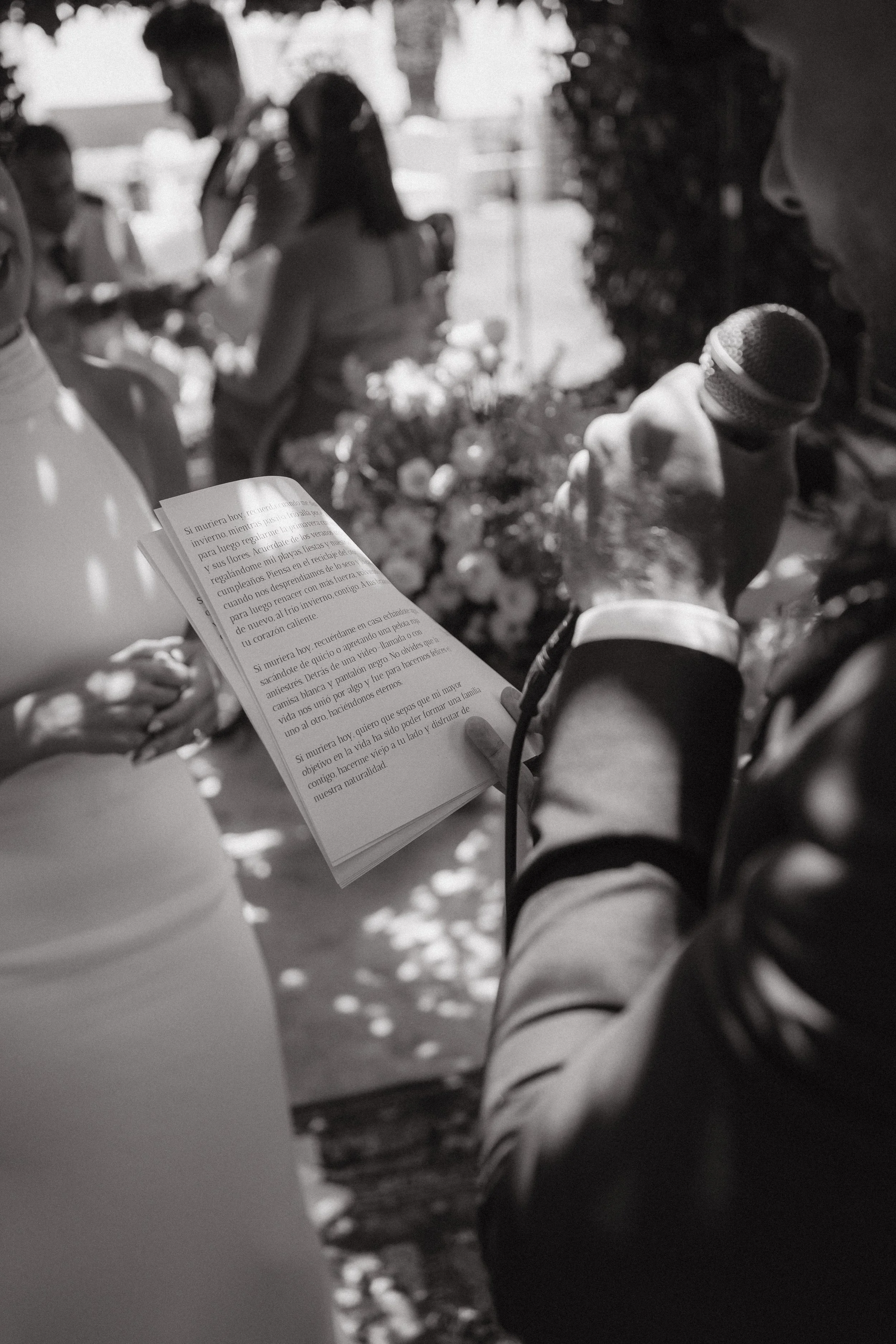 Un hombre vestido con traje, perfil visible, sosteniendo un micrófono y un libro abierto en una ceremonia al aire libre. Al fondo, varias personas, algunas con velos y vestimenta formal, participando en el evento. La escena está en blanco y negro.