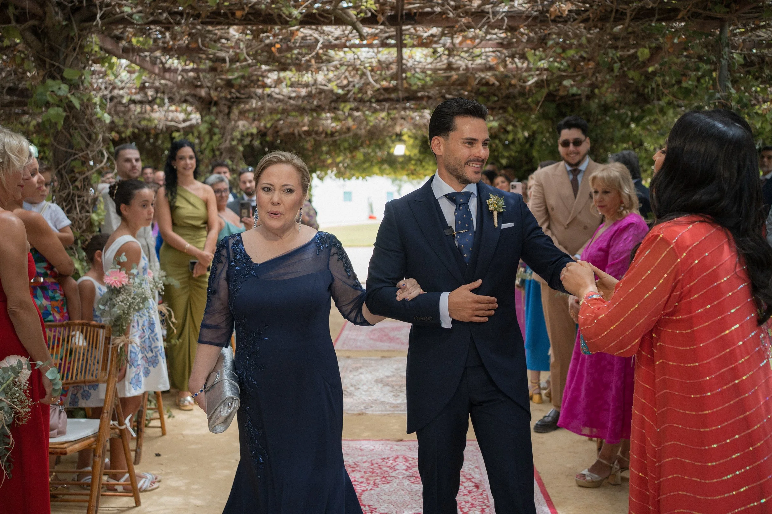 Entrada del novio con su madre, con invitados en el fondo, en un escenario decorado con enredaderas y flores.