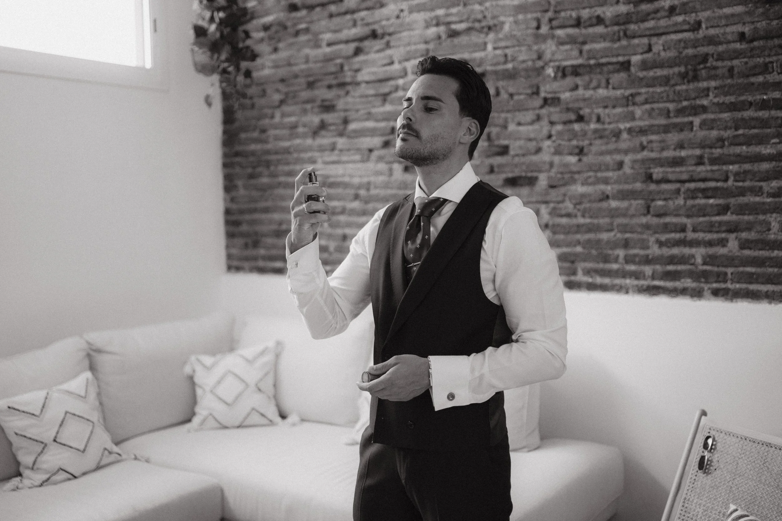Hombre con traje y corbata rociándose con un perfume en una sala con sofá y almohadas. En blanco y negro.