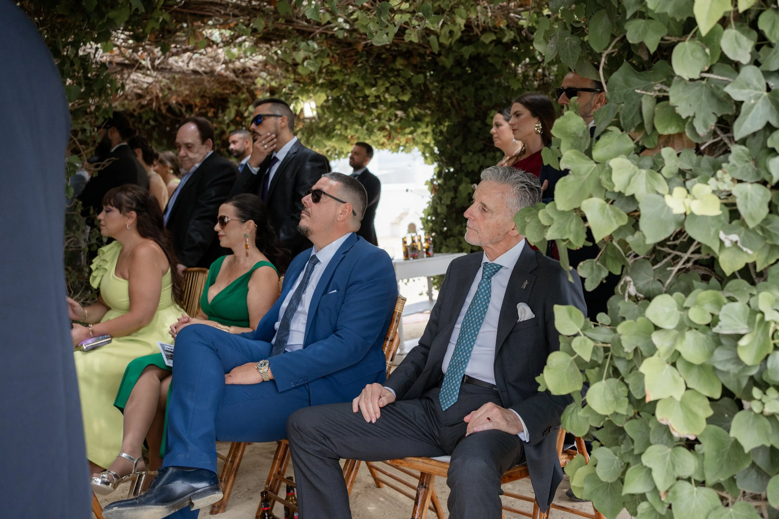 Personas sentadas y de pie en una ceremonia al aire libre, algunos con trajes formales, en un entorno con vegetación.