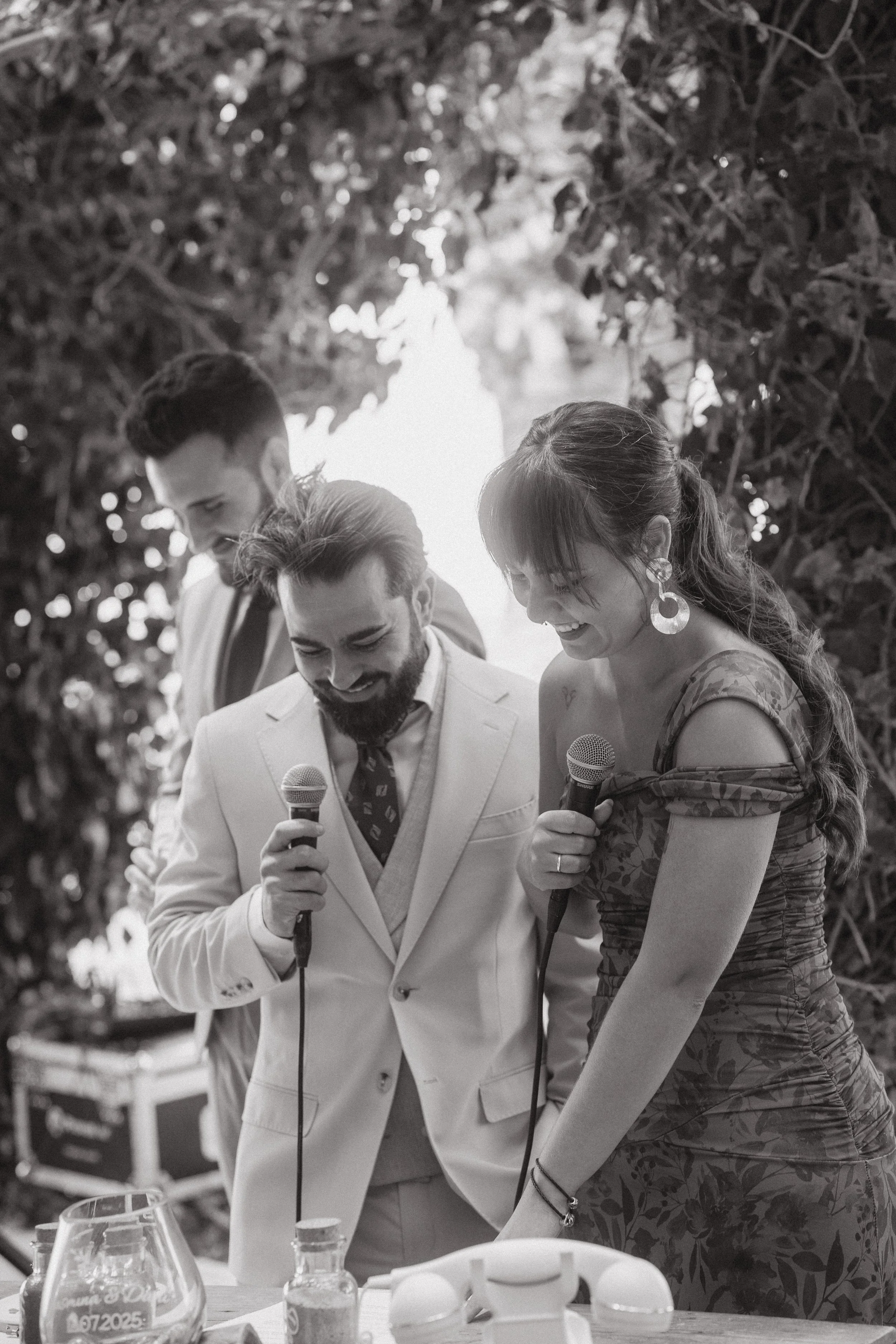 Tres personas en una reunión o celebración, sonriendo y sosteniendo micrófonos, en un entorno con arbustos o vegetación.