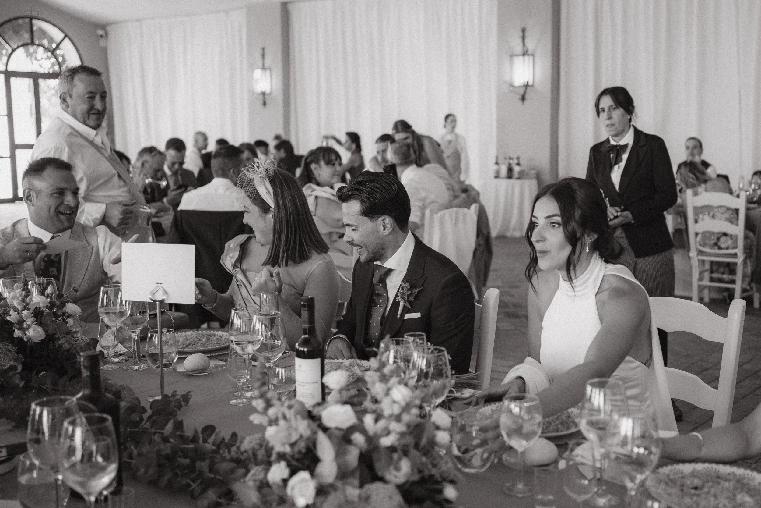 Personas celebrando en una boda, sentadas en una mesa decorada con flores, vino y comida, en un salón con cortinas blancas y lámparas