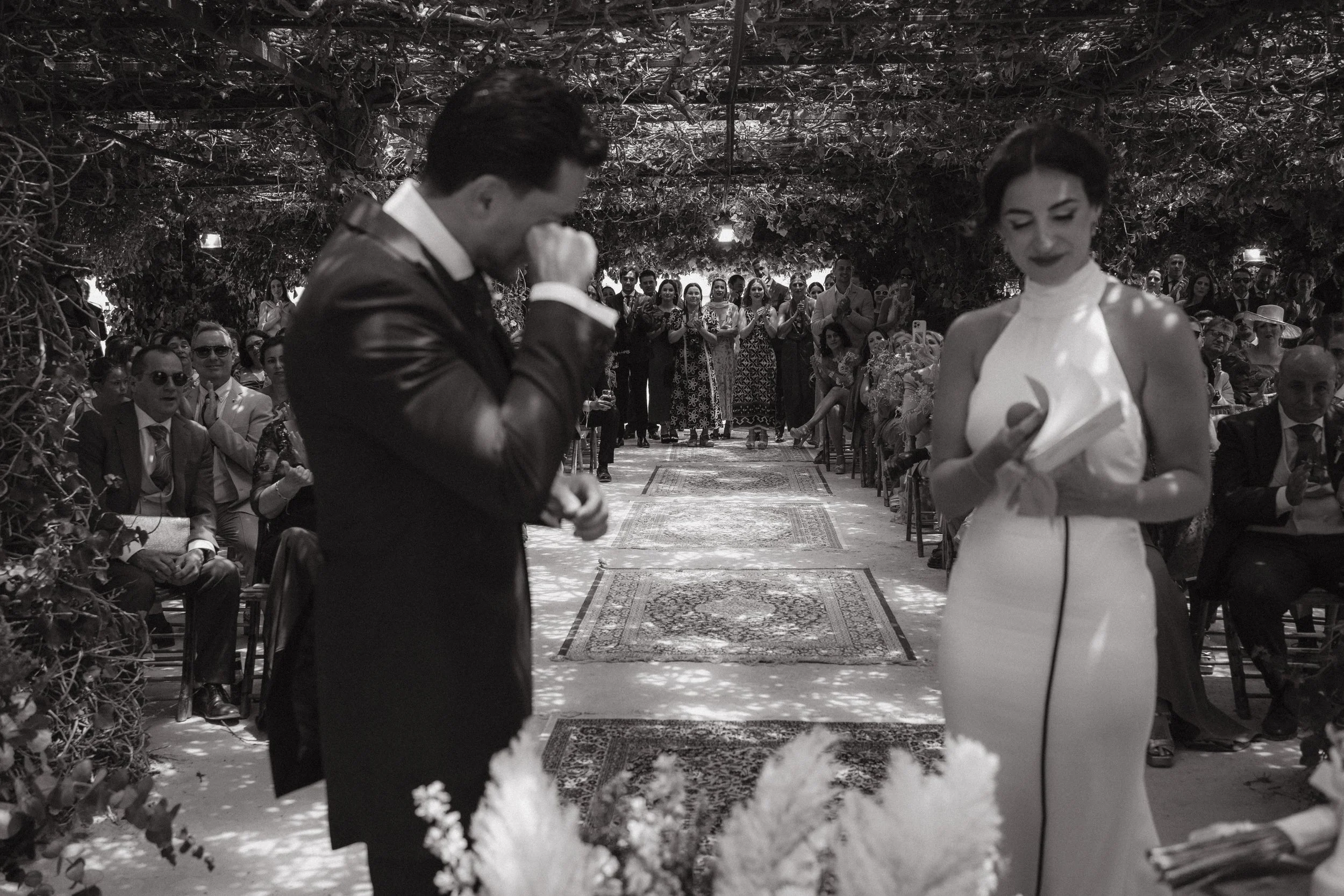 Una pareja en la ceremonia de matrimonio, la mujer sostiene una biblia y el hombre llora con la cabeza inclinada, en un lugar decorado con plantas y alfombras, rodeados de invitados sentados y observando.