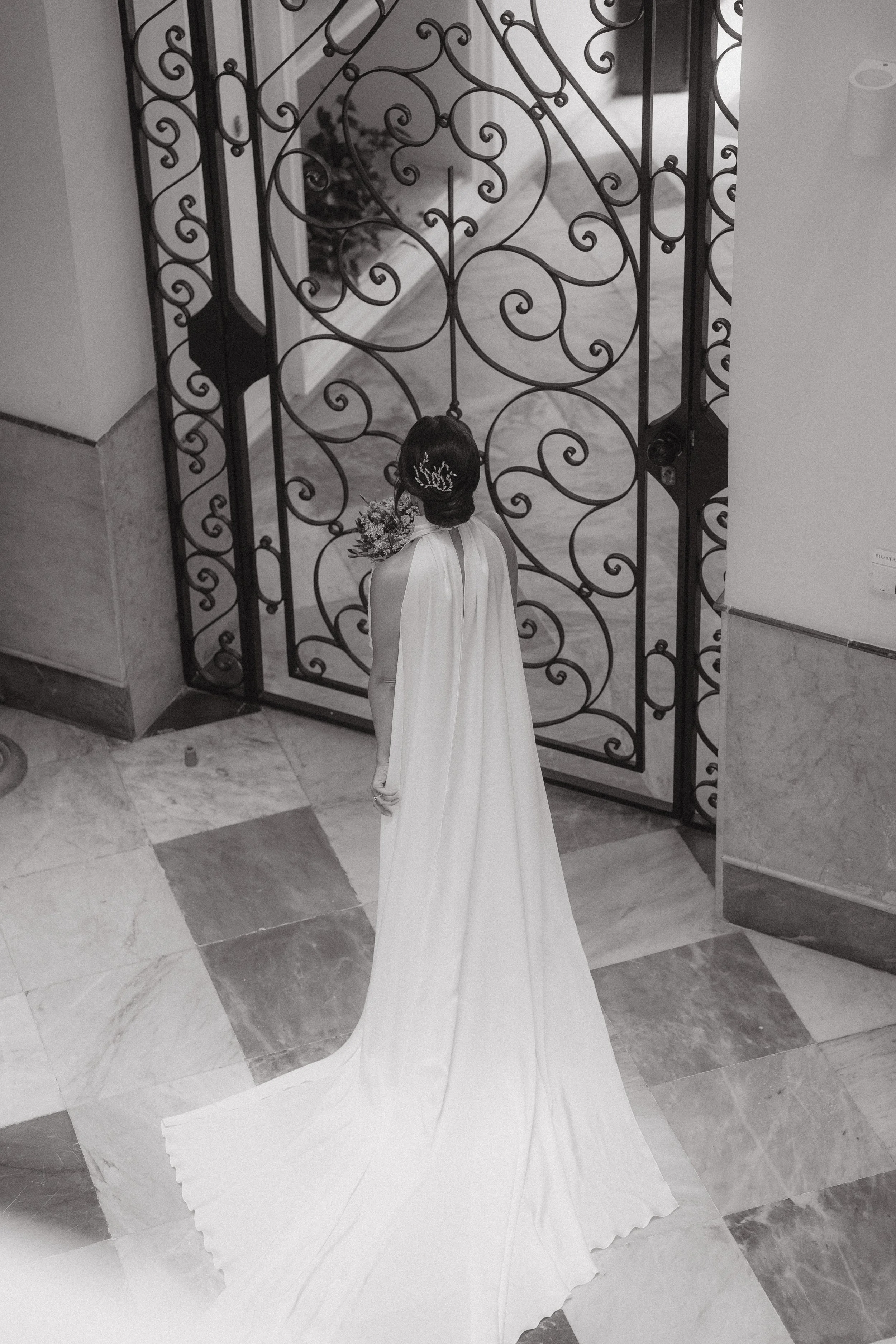 Mujer con vestido de novia y tiara, de espaldas, frente a una puerta de hierro con detalles decorativos.