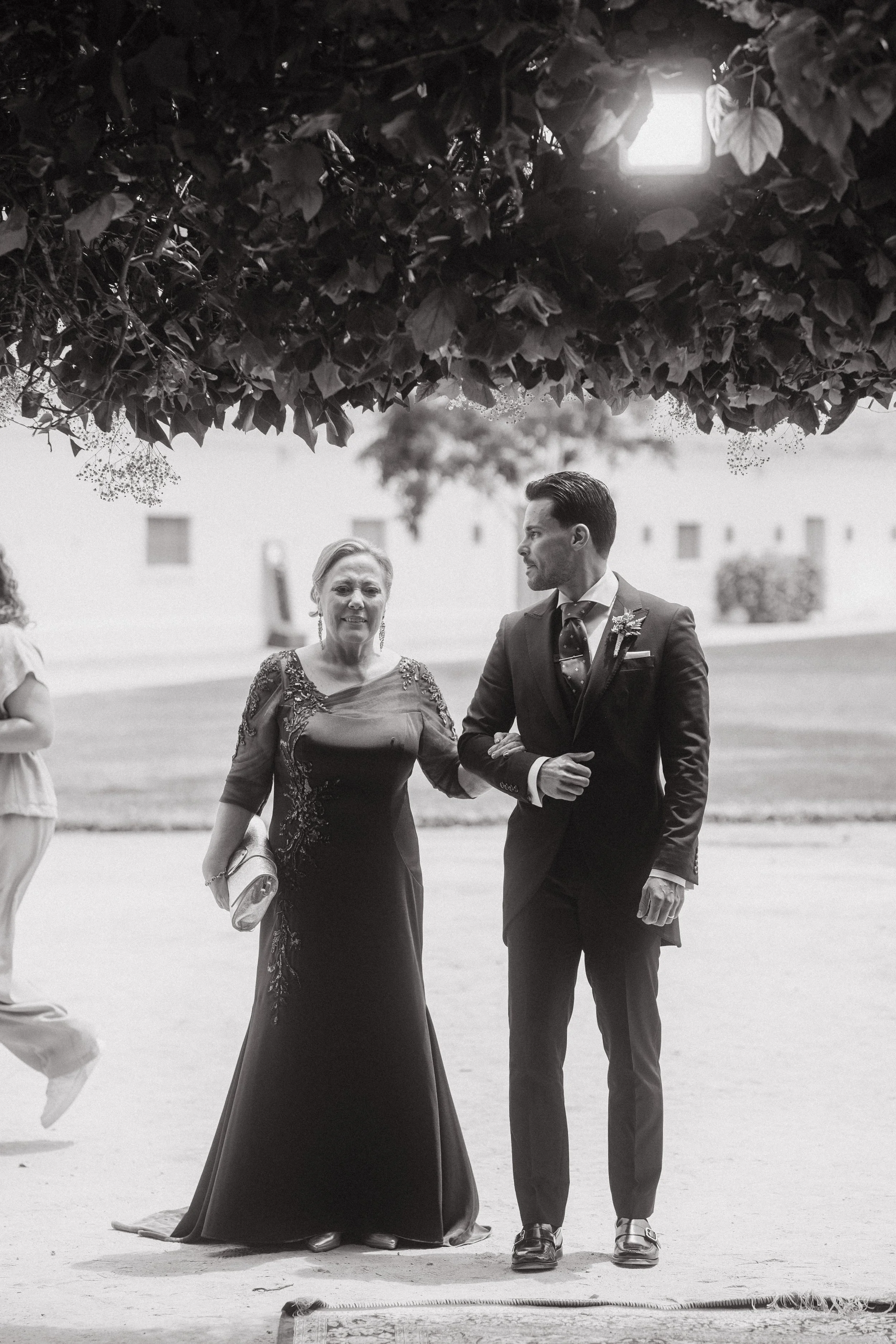 Una mujer mayor vestida con un vestido largo y un hombre joven con traje caminando juntos bajo un árbol