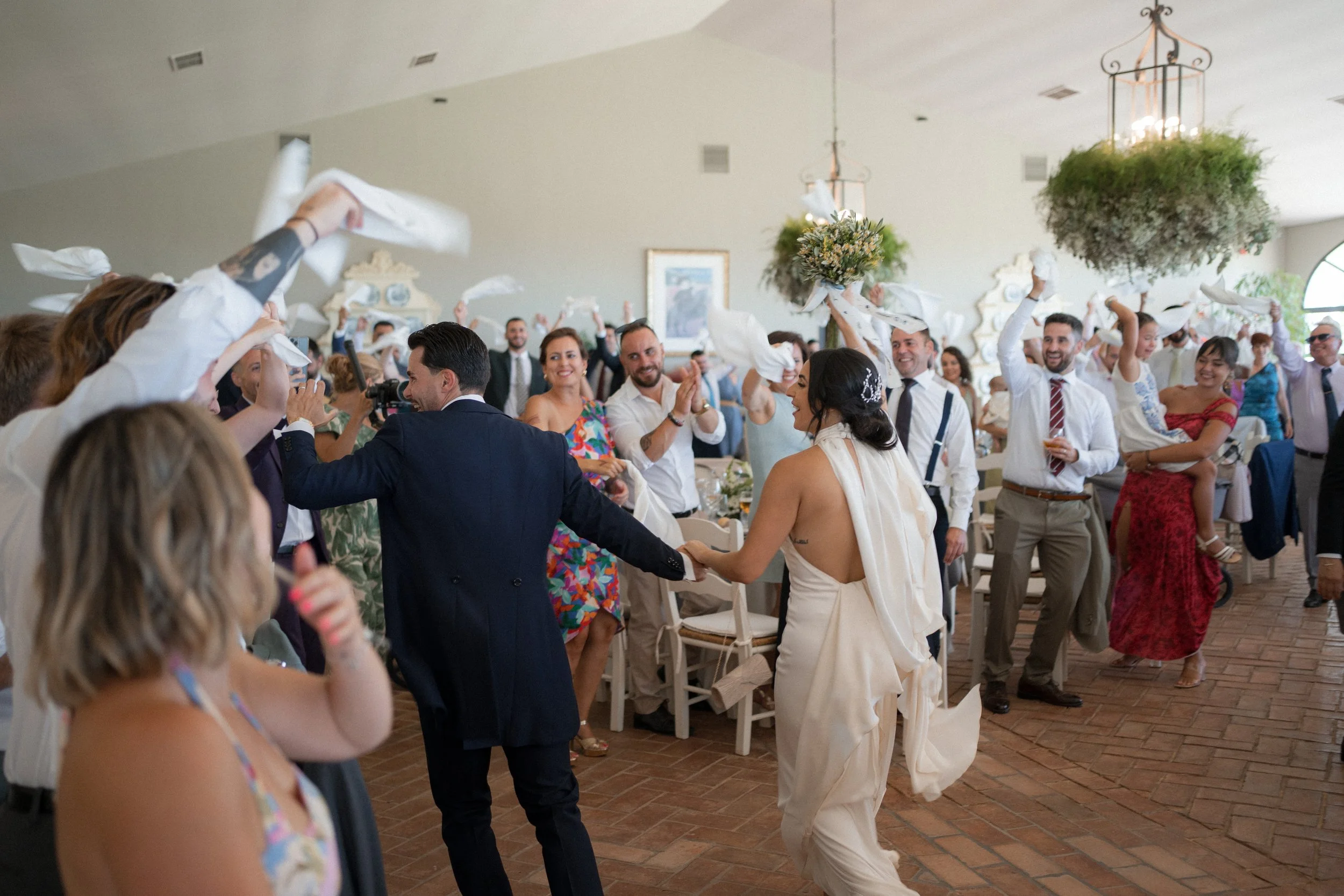 Pareja de novios bailando con invitados en una celebración de boda en un salón decorado con plantas y candelabros.