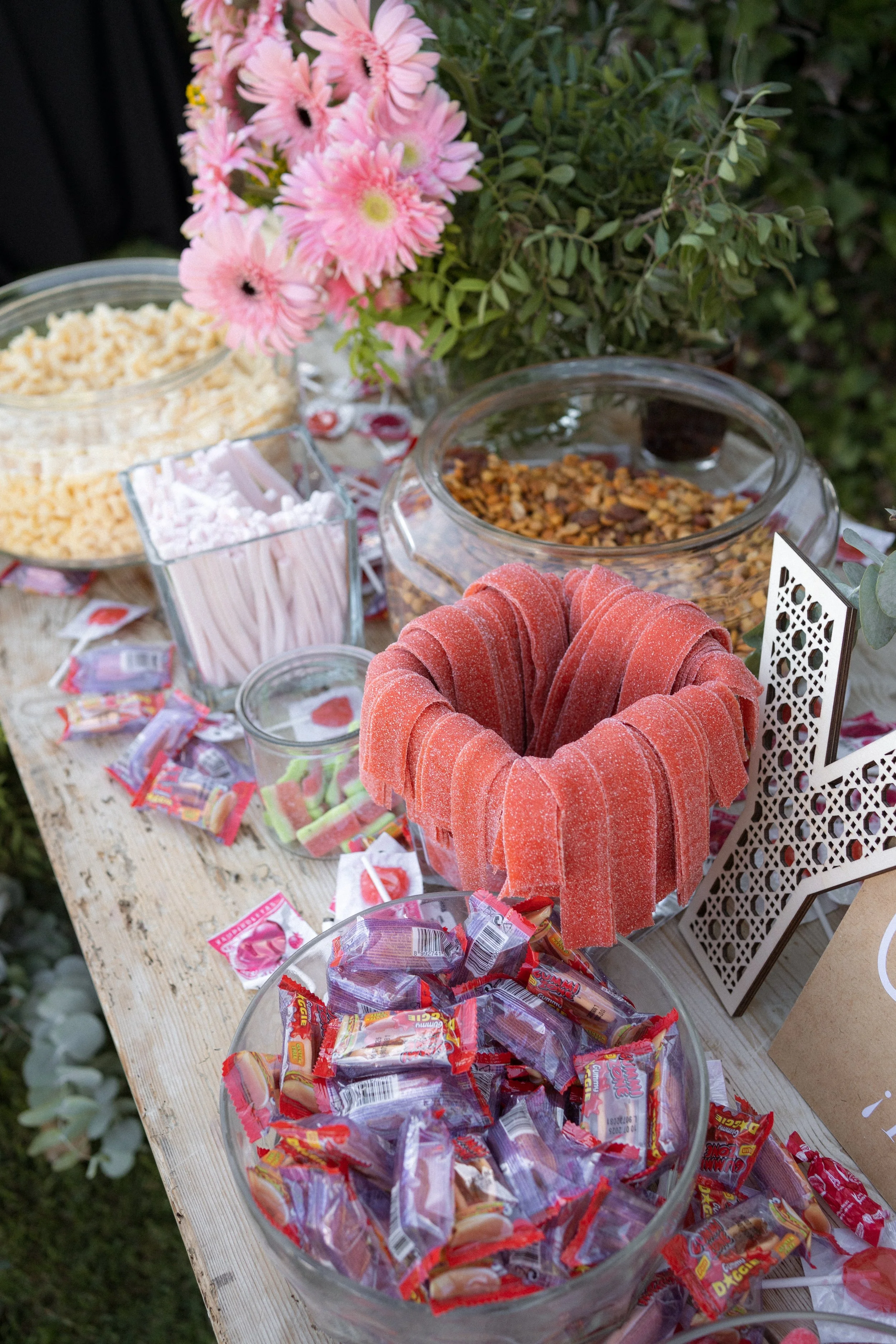 Mesa con dulces y caramelos, jarras con confites, malvaviscos, y una corona de gomitas rojas, decorada con flores rosadas y verdes.