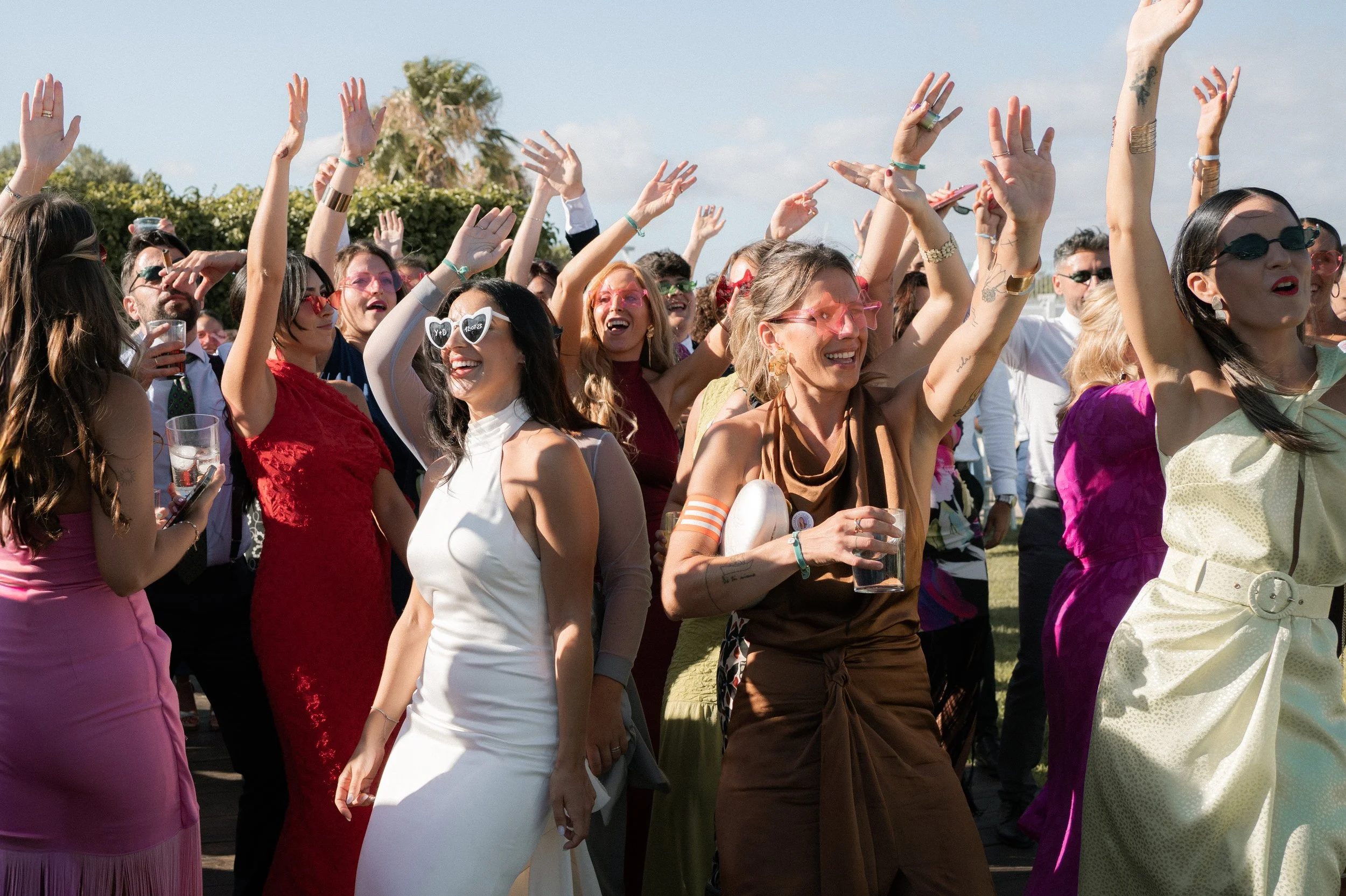 Grupo de personas disfrutando en una fiesta al aire libre, bailando y levantando las manos, con expresión de alegría y sonrisas, en un día soleado.