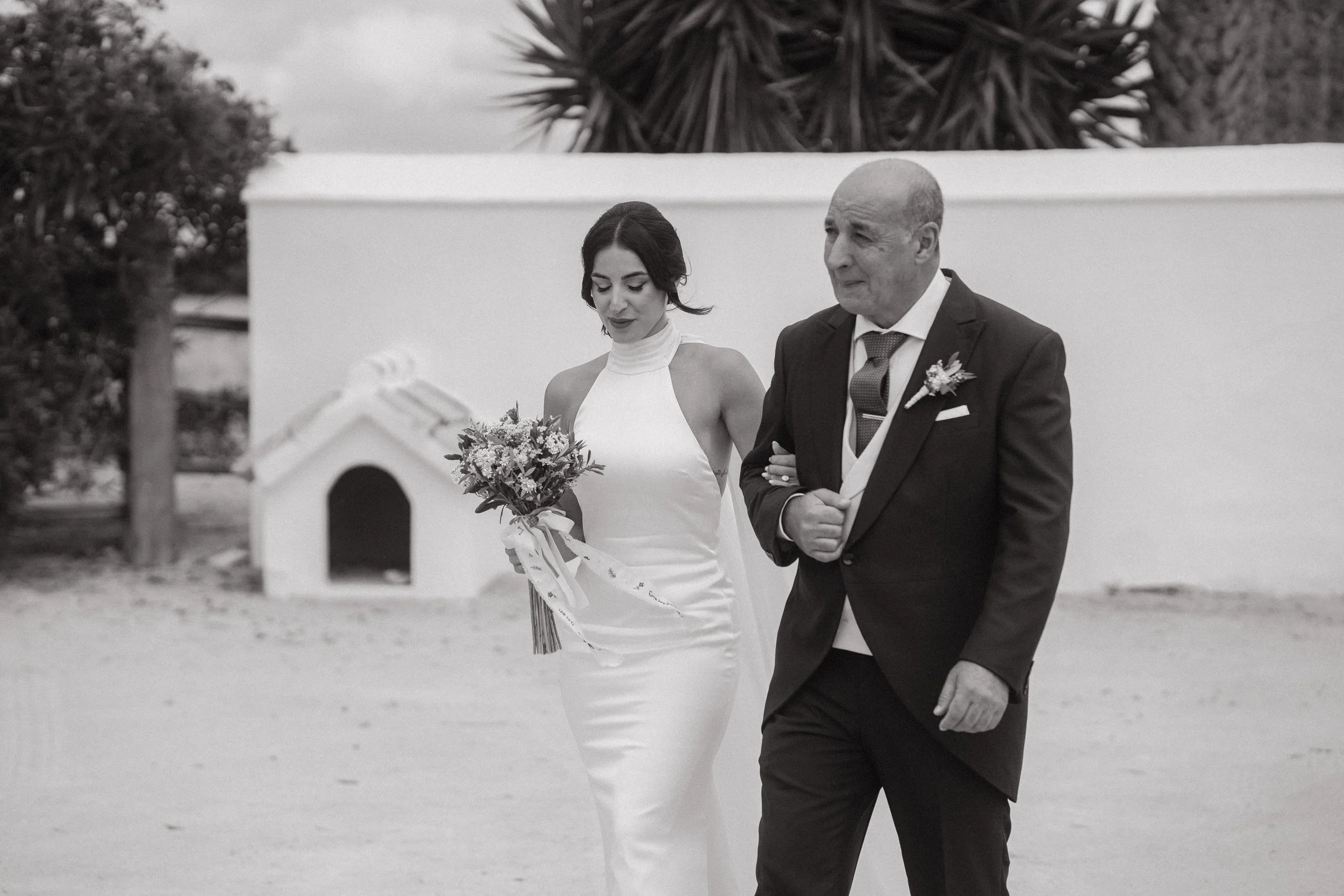 Una novia con vestido blanco y un hombre con traje de boda caminando juntos, en un entorno exterior, en blanco y negro.