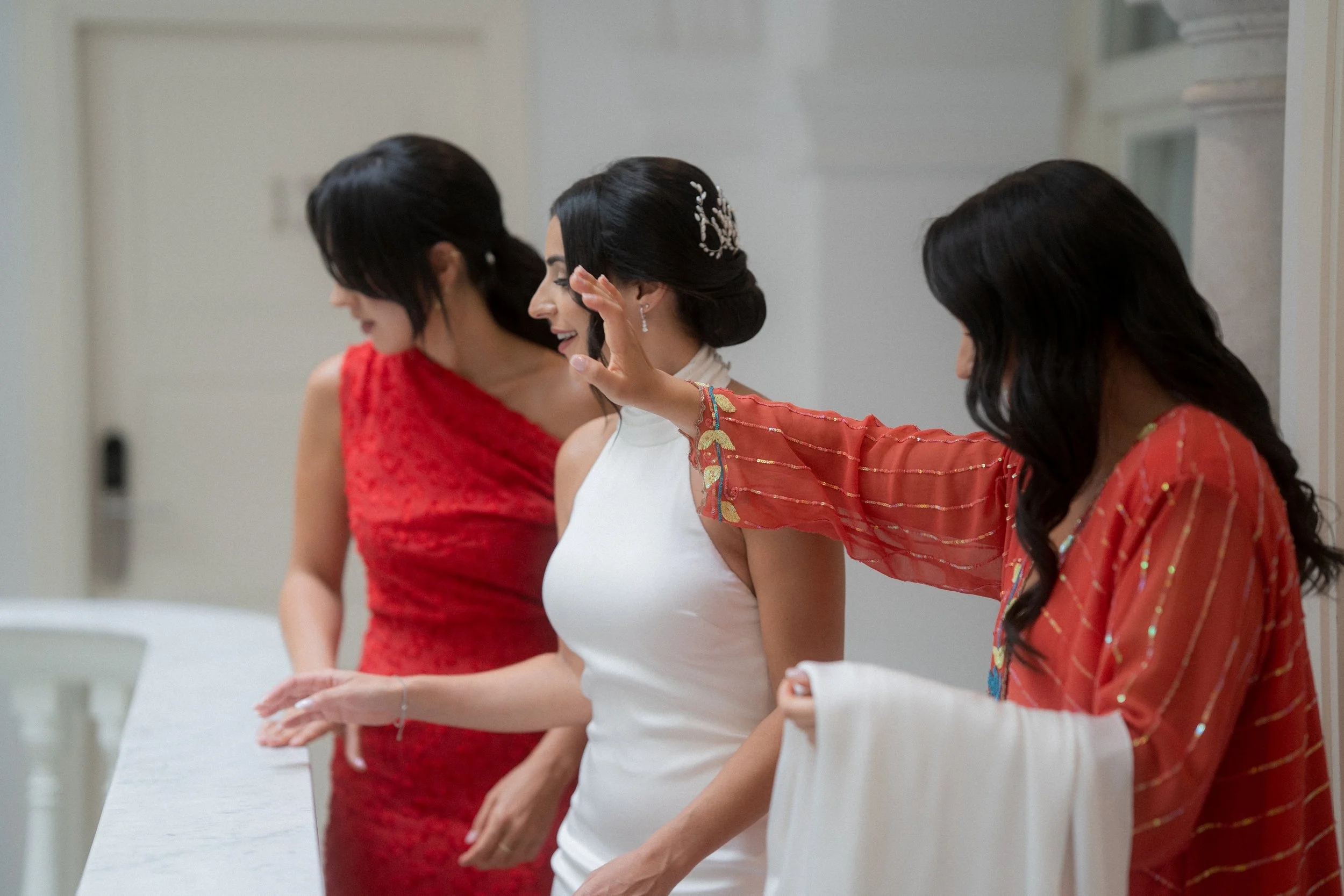 Tres mujeres en vestido de boda tienen un momento de alegría, una de ellas con vestido blanco y otra con vestido rojo.