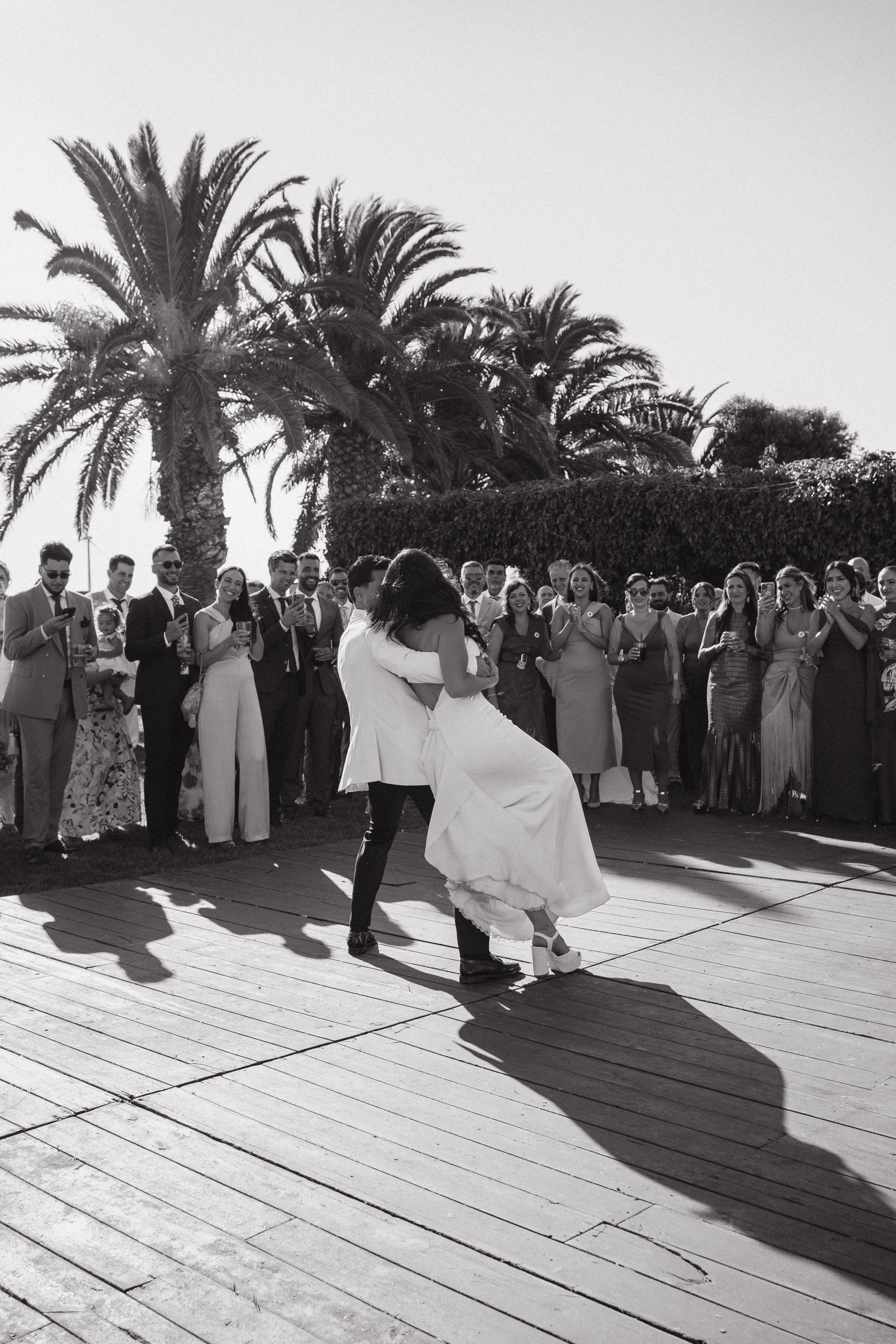 Pareja bailando en una ceremonia al aire libre, rodeada de invitados y palmeras en el fondo.