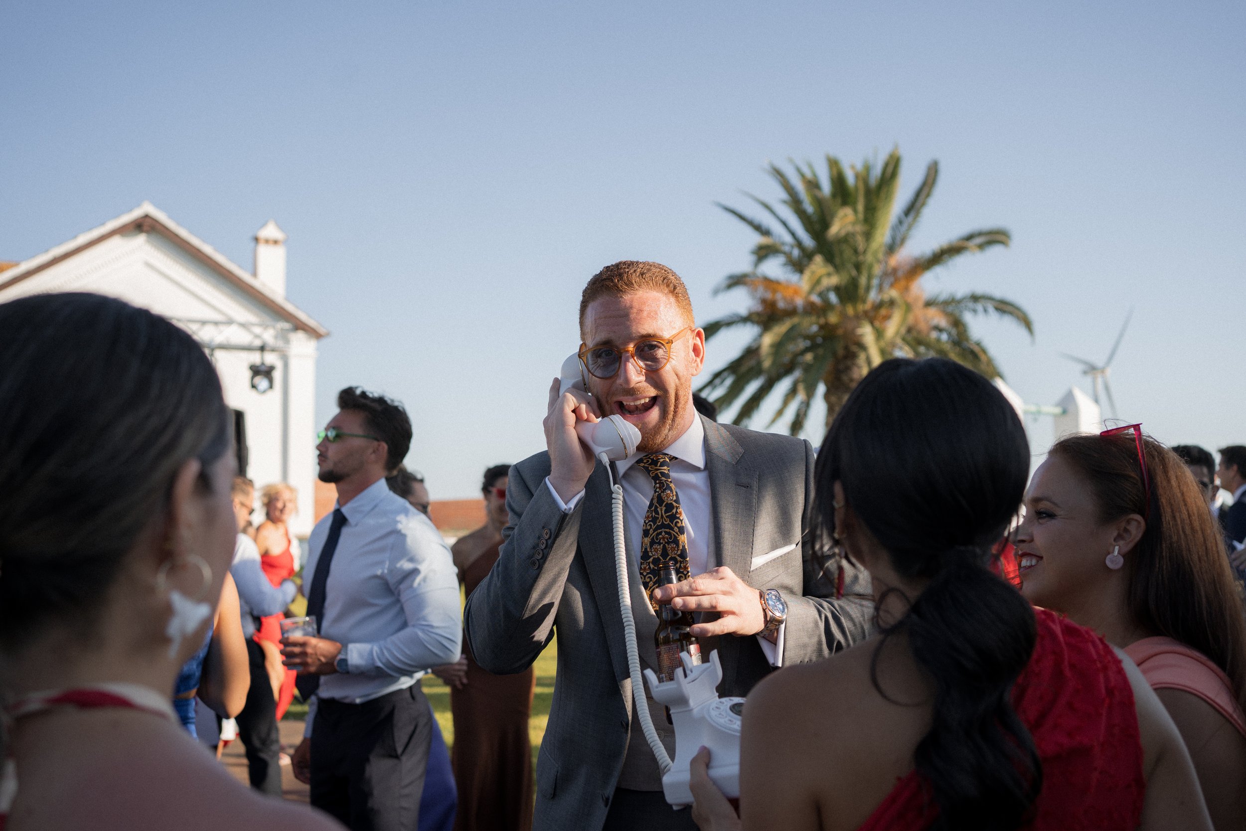 Hombre con gafas y traje gris hablando por teléfono en una reunión social al aire libre con otras personas y palmeras al fondo.