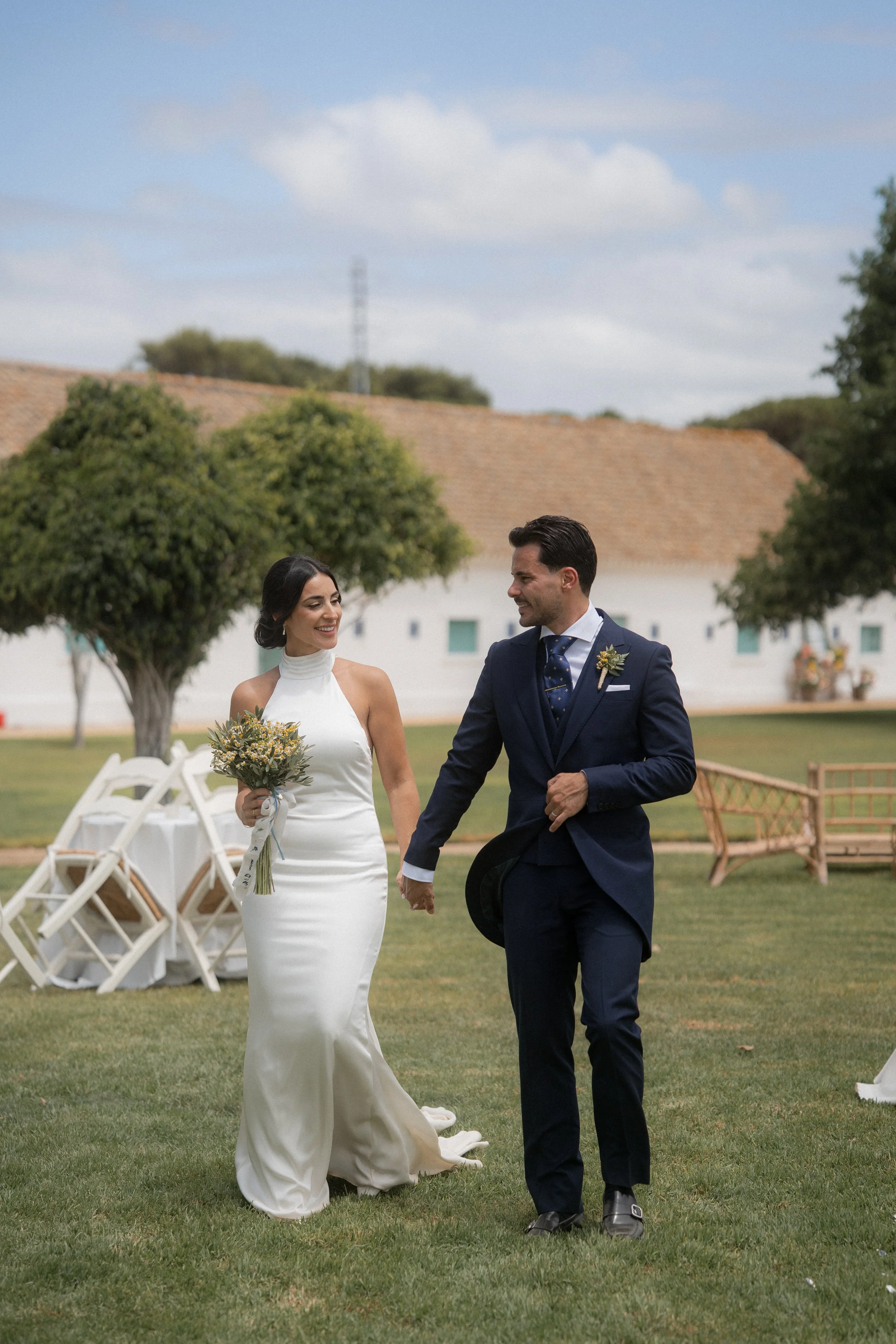 Pareja en boda, la mujer con vestido de novia y el hombre con traje formal, caminando en un jardín durante el día.