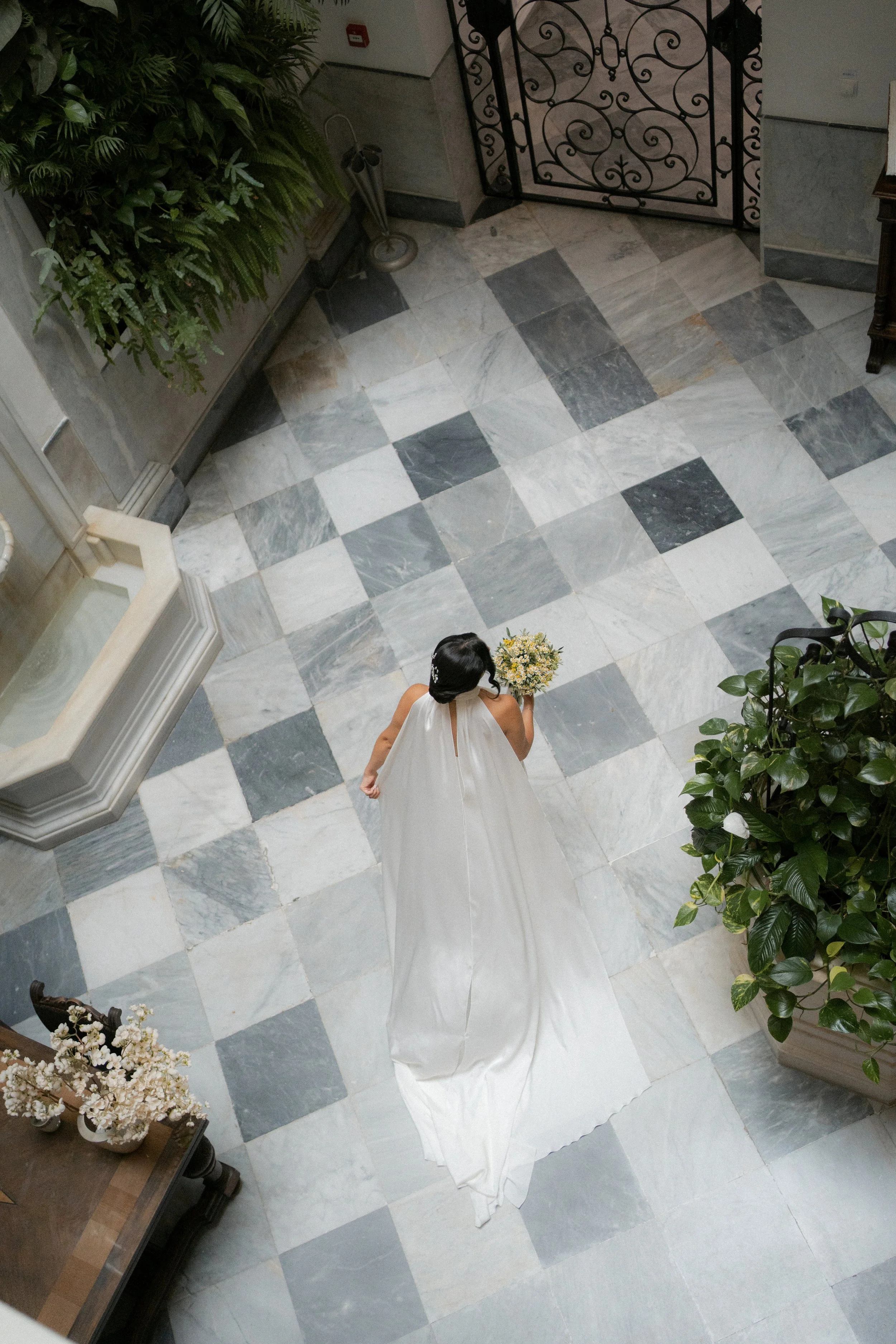 Mujer con vestido de novia caminando por un pasillo con pisos de mármol, sosteniendo un ramo de flores.
