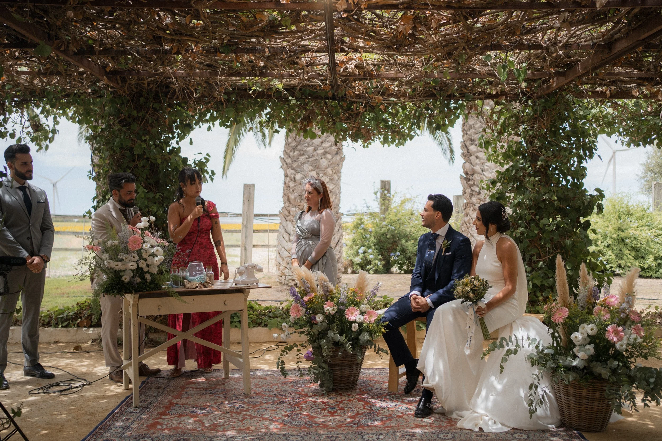 Una ceremonia de boda al aire libre con una pareja sentada y varias personas de pie, decoraciones florales, bajo un arco de madera cubierto de plantas y flores.