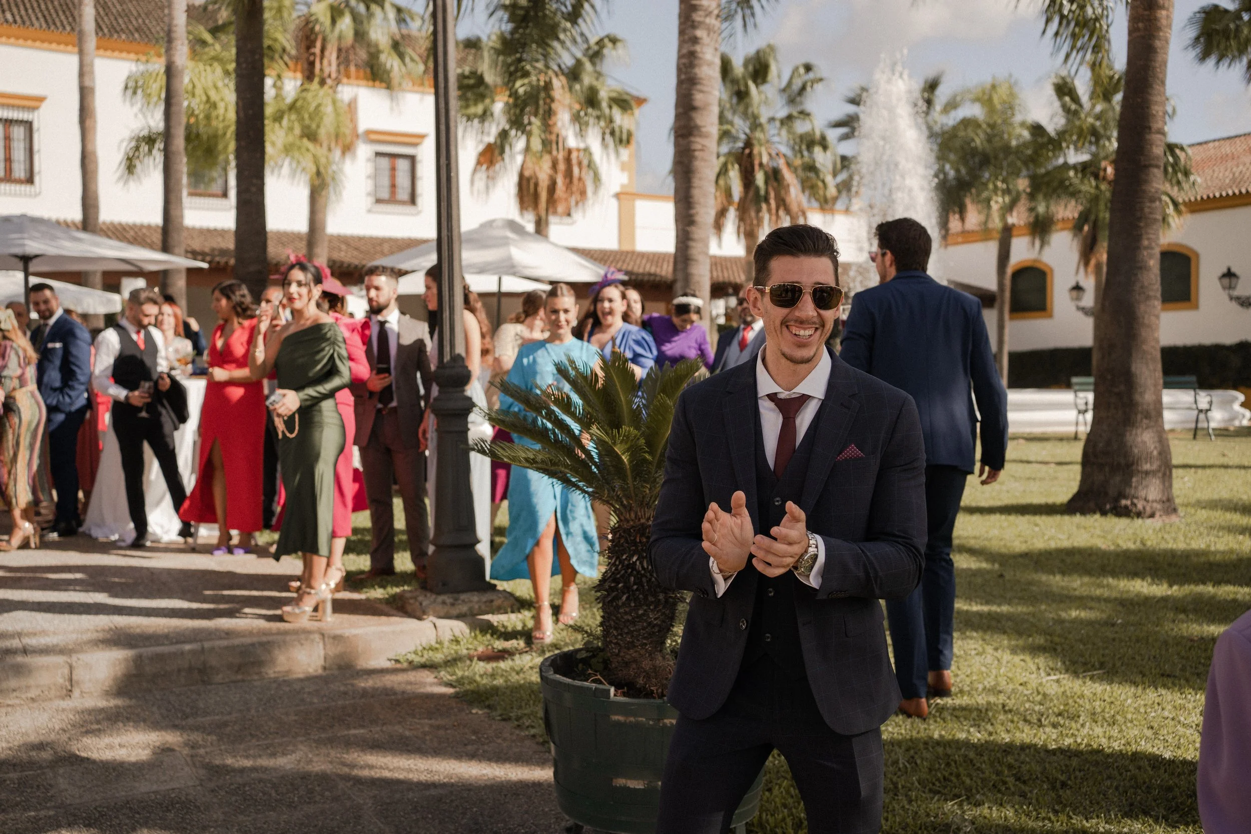 Grupo de personas bien vestidas en un evento al aire libre con palmeras y una fuente de fondo.