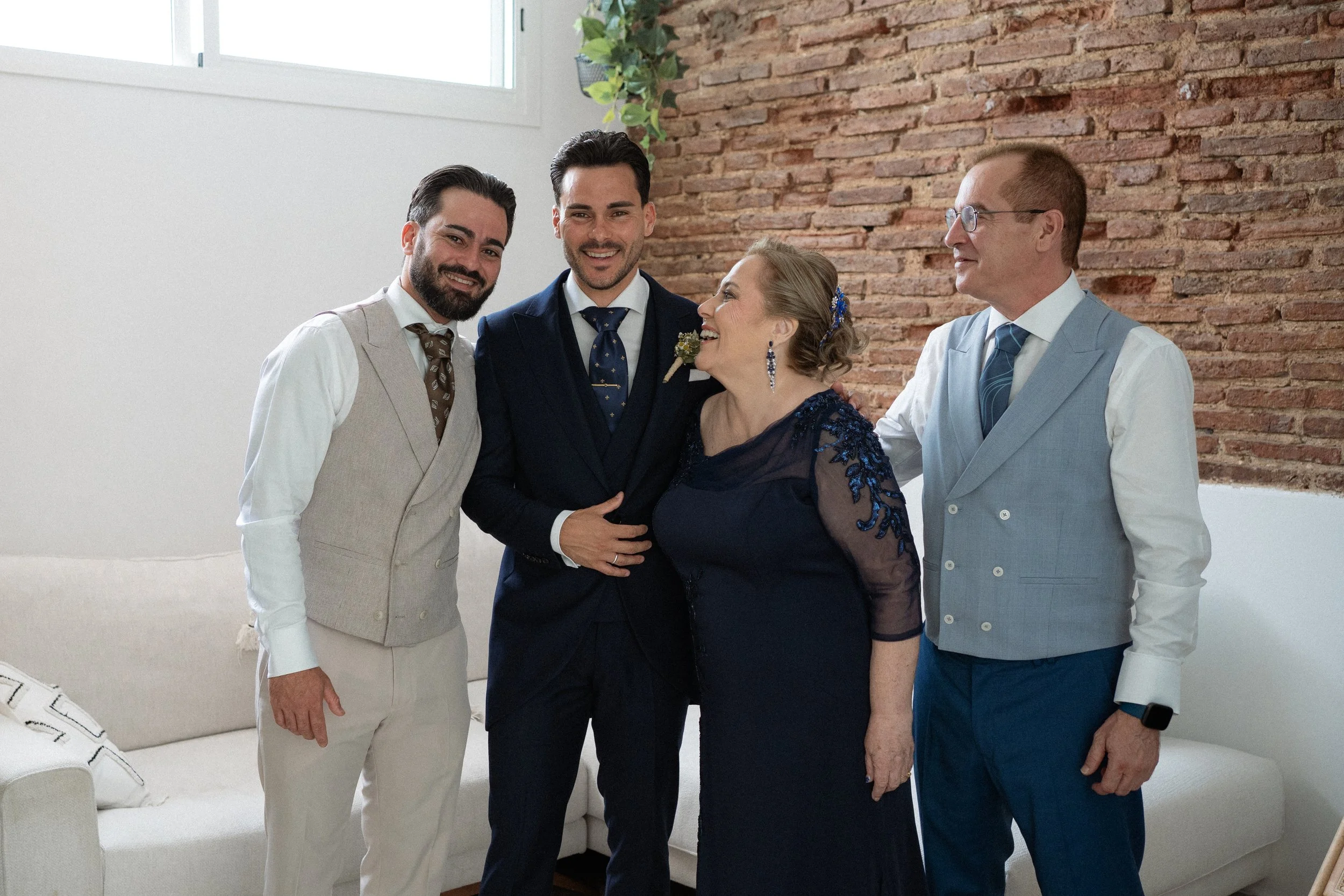 Cuatro personas en traje formal en una reunión familiar o celebración, con paredes de ladrillo y un sofá beige en el fondo.