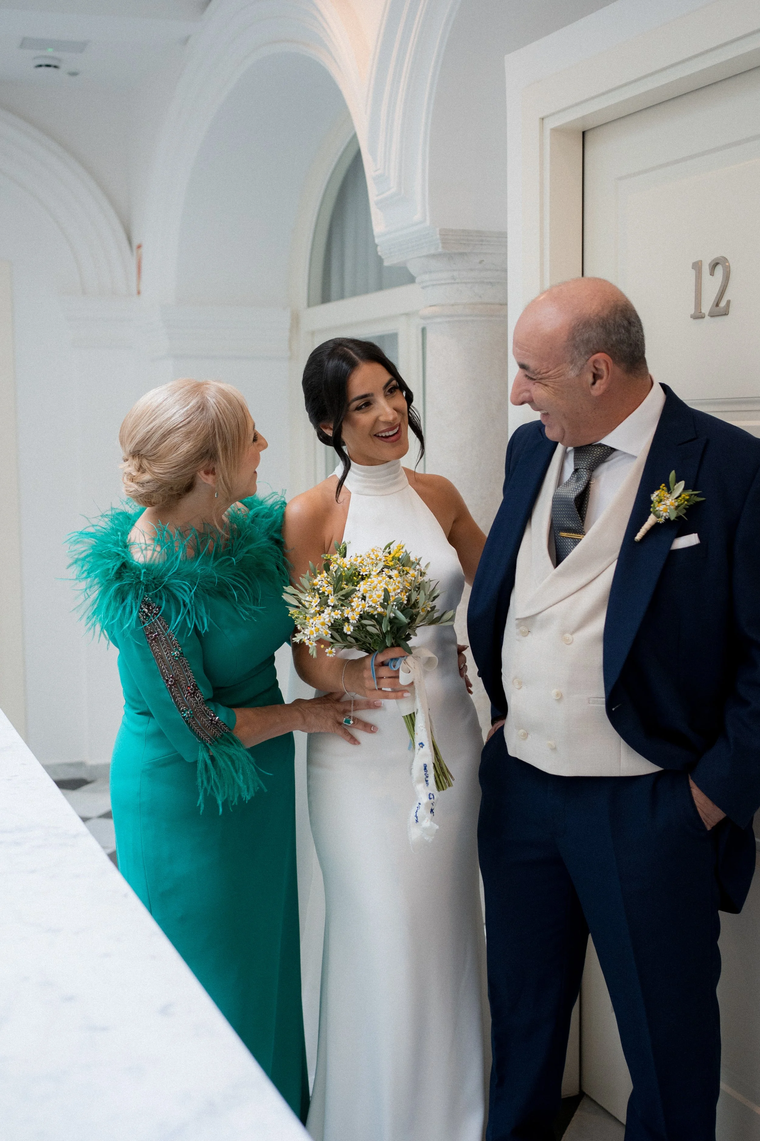 Mujer con vestido blanco sosteniendo un ramo de flores, rodeada de una mujer mayor con vestido verde y un hombre con traje de novio, en un vestíbulo elegante del hotel El armador en Cádiz.