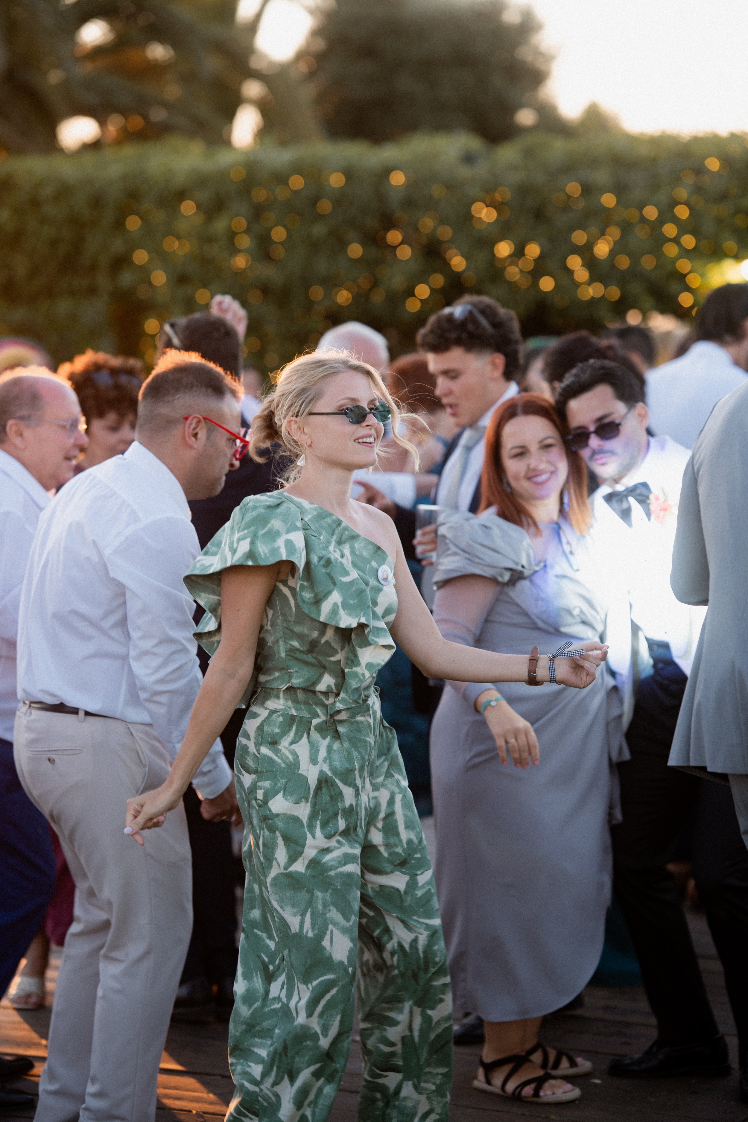 Personas bailando en una celebración al aire libre con luces decorativas y árboles de fondo.
