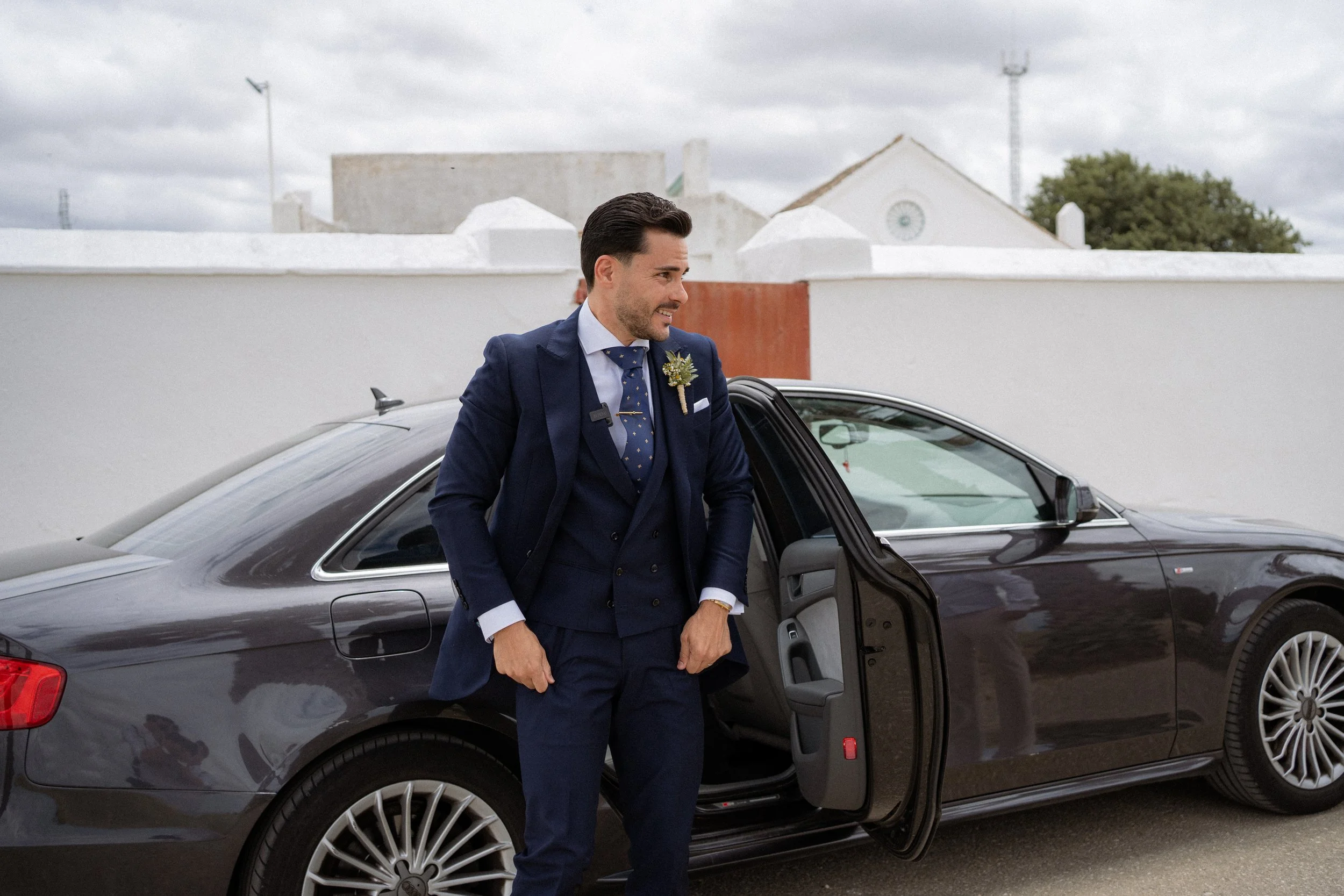 Hombre vestido en traje azul entrando a un coche negro, en un fondo de muro blanco y cielo nublado.