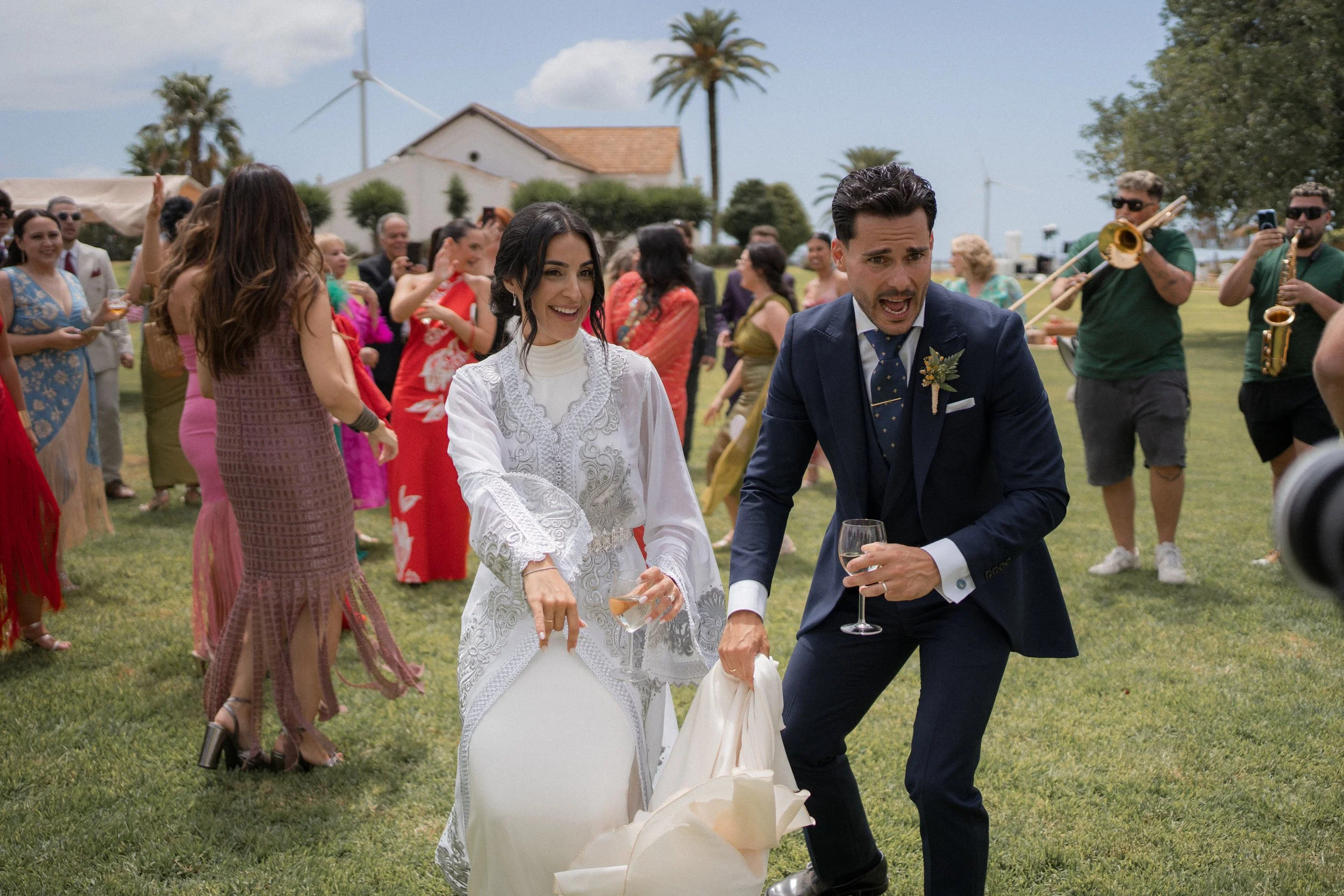 Pareja de recién casados bailando en una celebración al aire libre, rodeados de invitados y músicos, con un fondo de cielo soleado, palmeras y casas.