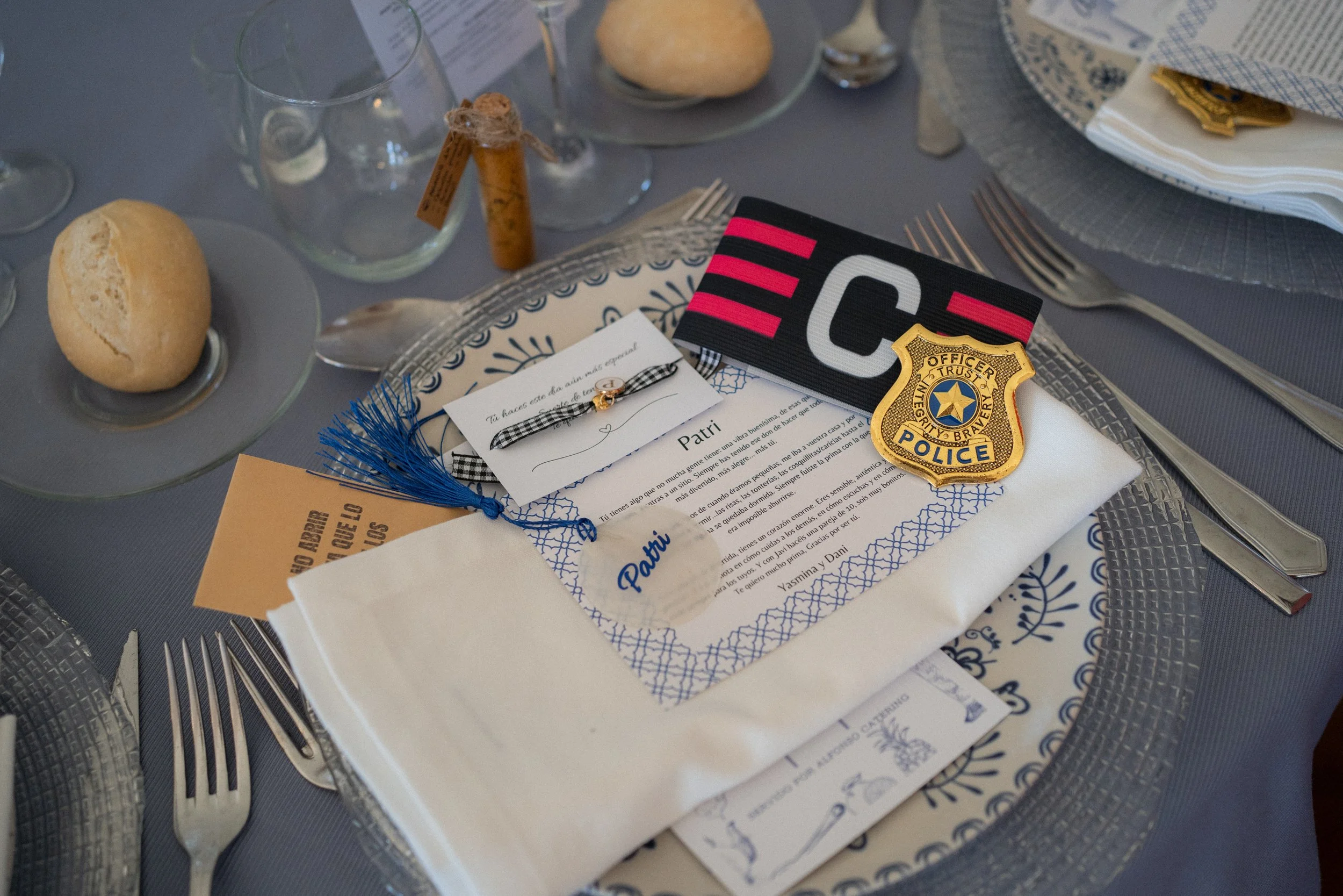Tarjeta de invitación decorada con un lazo, una medalla de policía, una base de vela, y otros accesorios, colocada en un plato en una mesa con cubiertos, cristalería y pan.