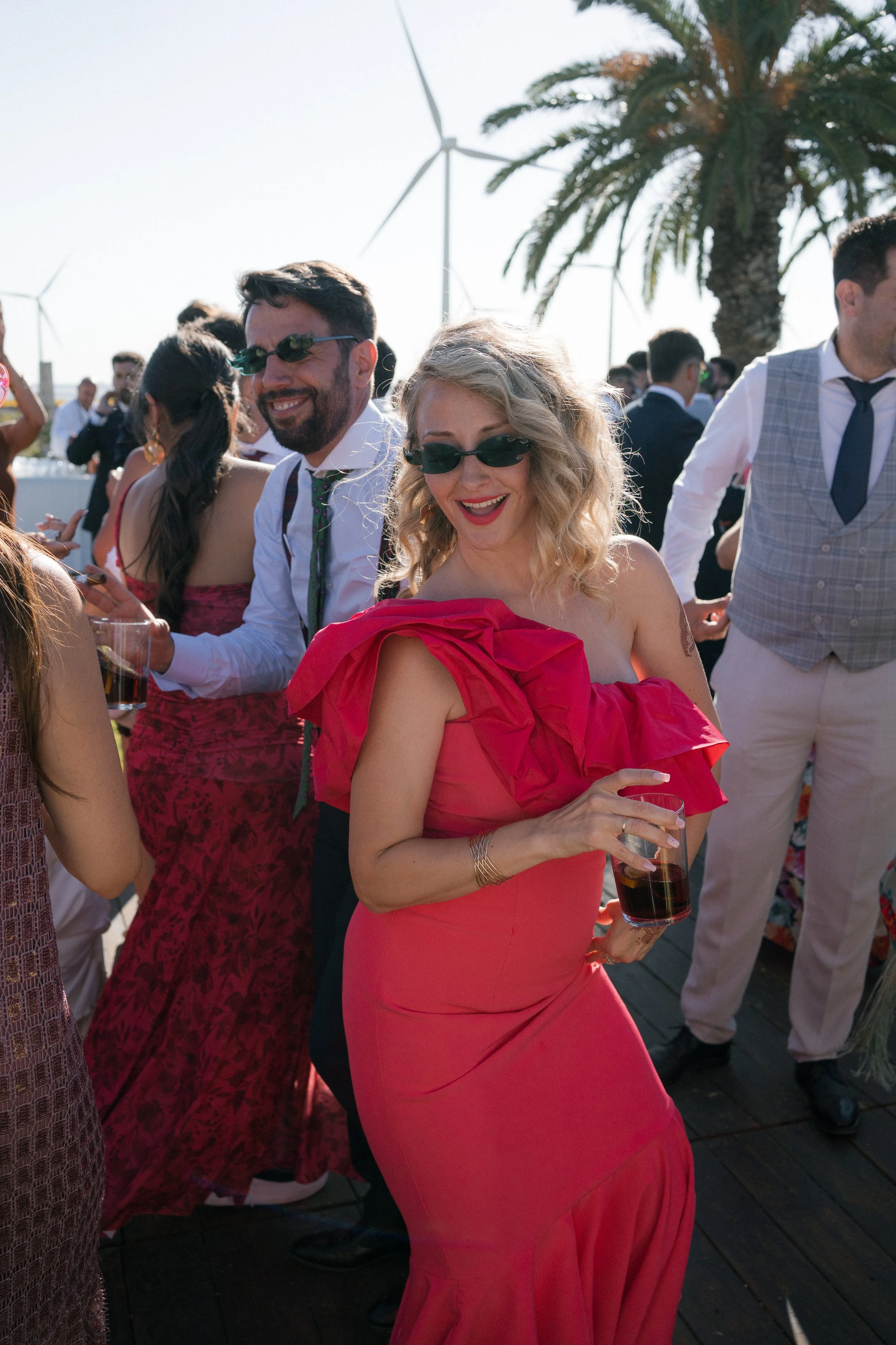 Grupo de personas disfrutando en una celebración al aire libre, con palmeras y molinos de viento en el fondo, una mujer en vestido rojo tomando una bebida y sonriendo.