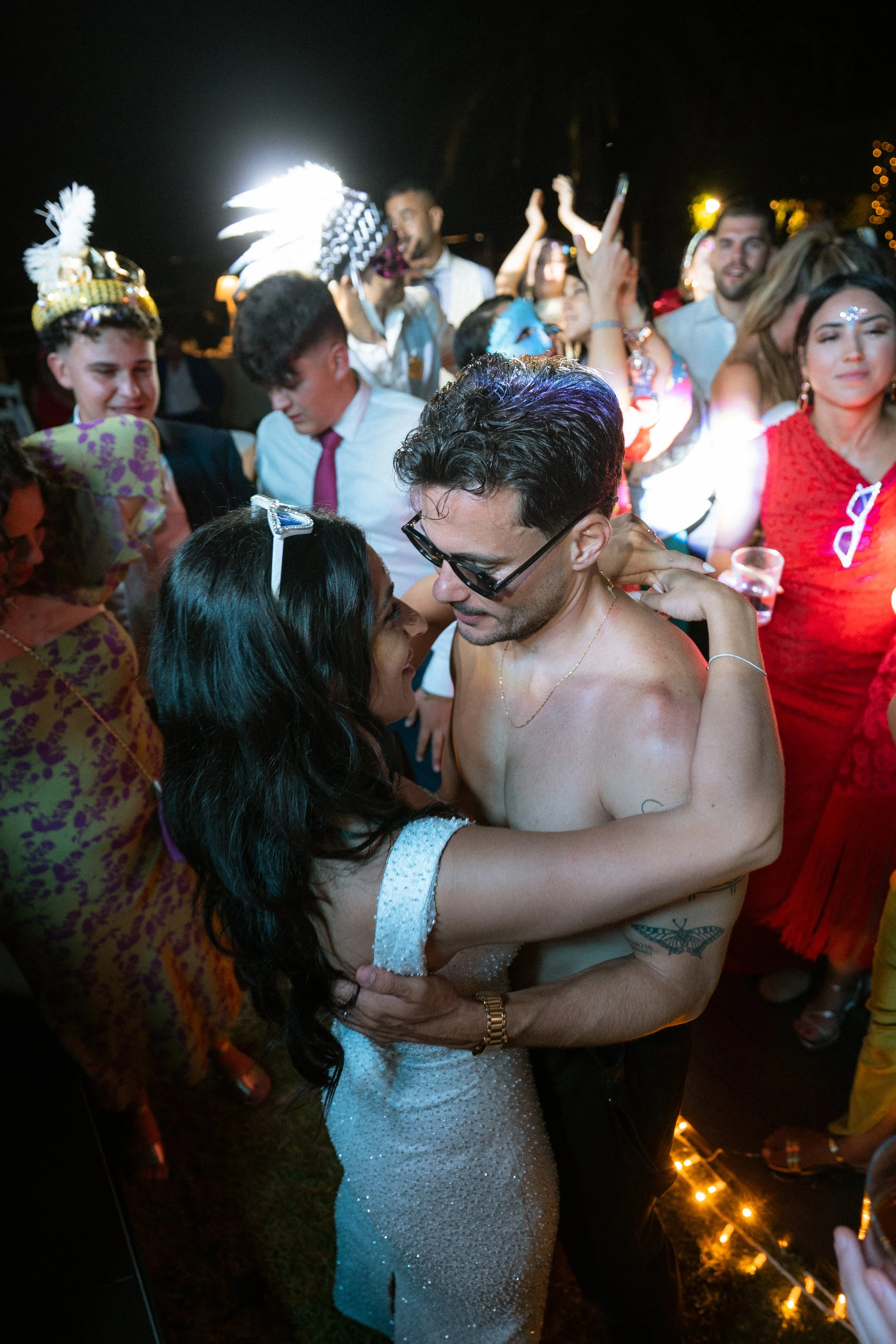 Pareja bailando en una fiesta, rodeada de amigos con disfraces y ropa colorida, en un ambiente festivo nocturno.