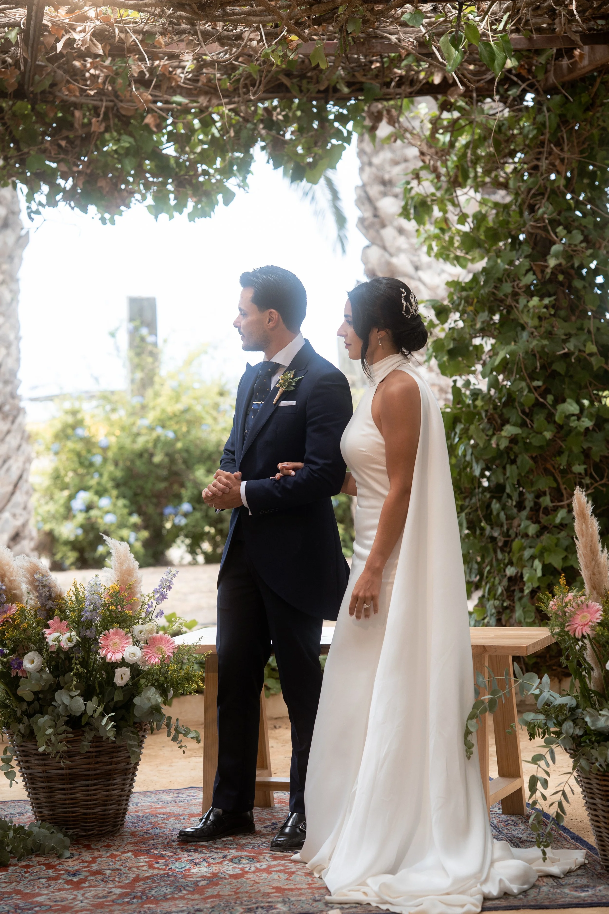 Una pareja en una ceremonia de boda, la mujer lleva un vestido blanco y el hombre un traje oscuro, están juntos bajo un arco decorado con plantas y flores.
