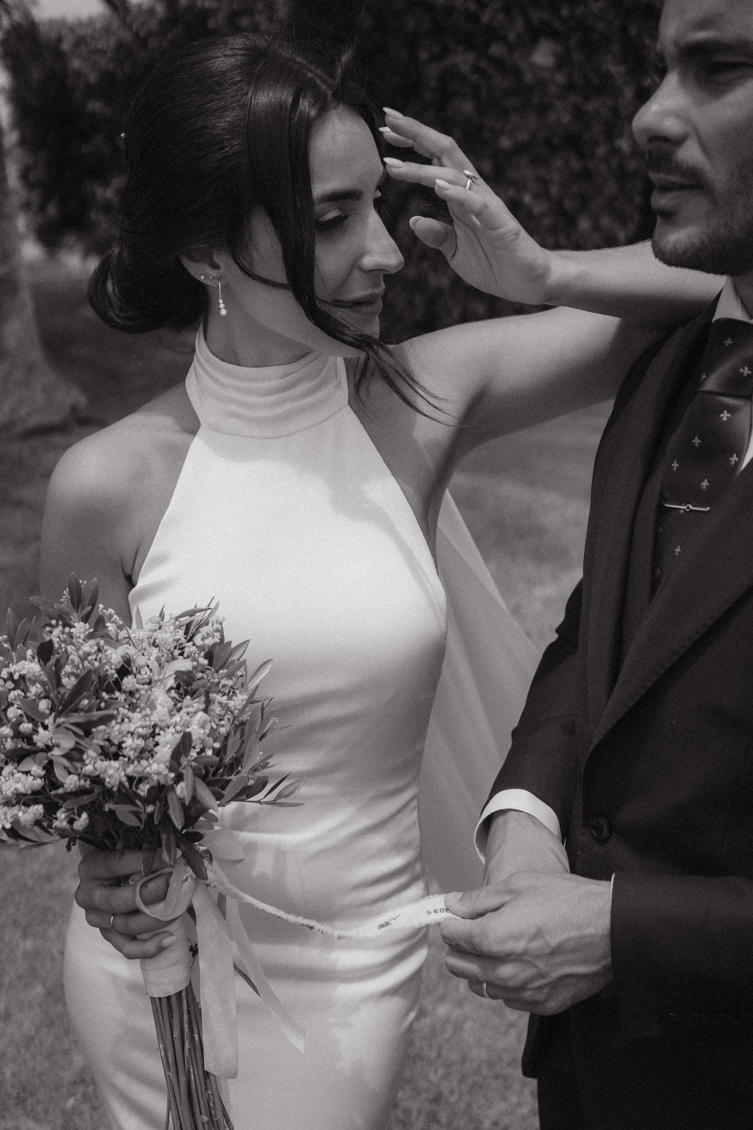 Una pareja en una ceremonia de boda, la mujer sostiene un ramo de flores y el hombre la sostiene de la mano, ambos parecen estar en un momento emotivo.