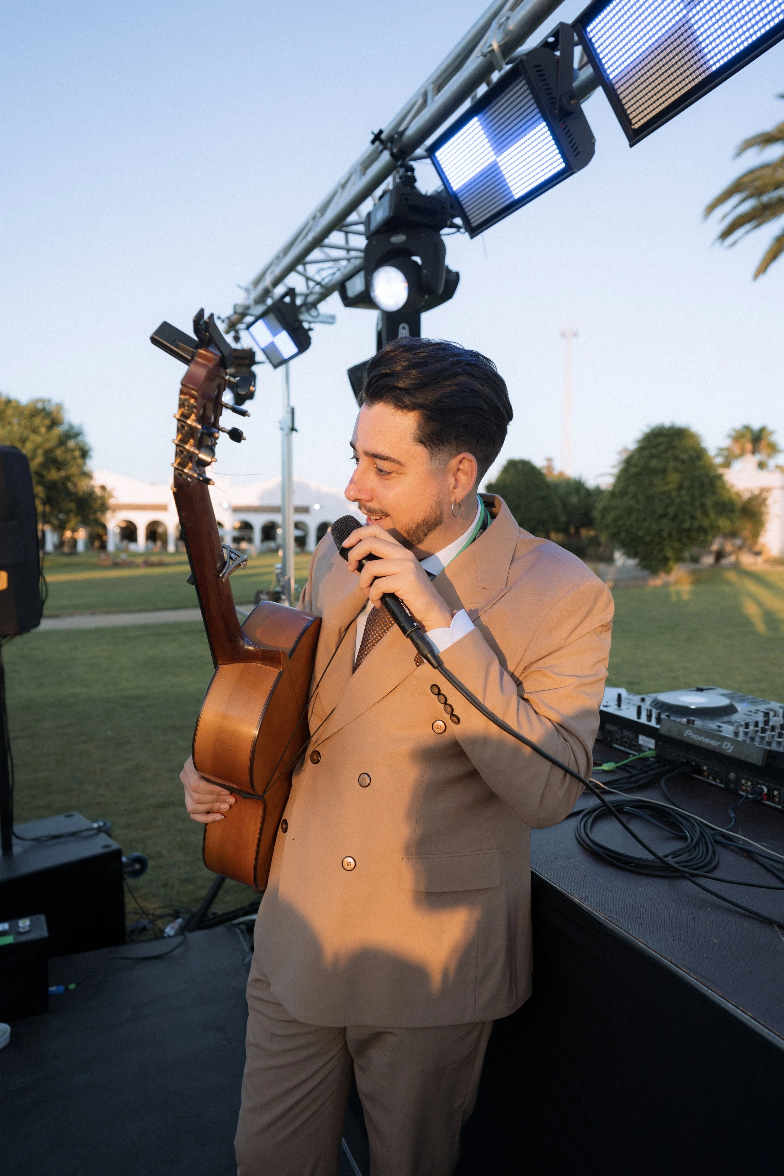 Hombre con un traje beige tocando la guitarra acústica y cantando en un micrófono, en un escenario al aire libre con equipo de música y luces, durante el atardecer.