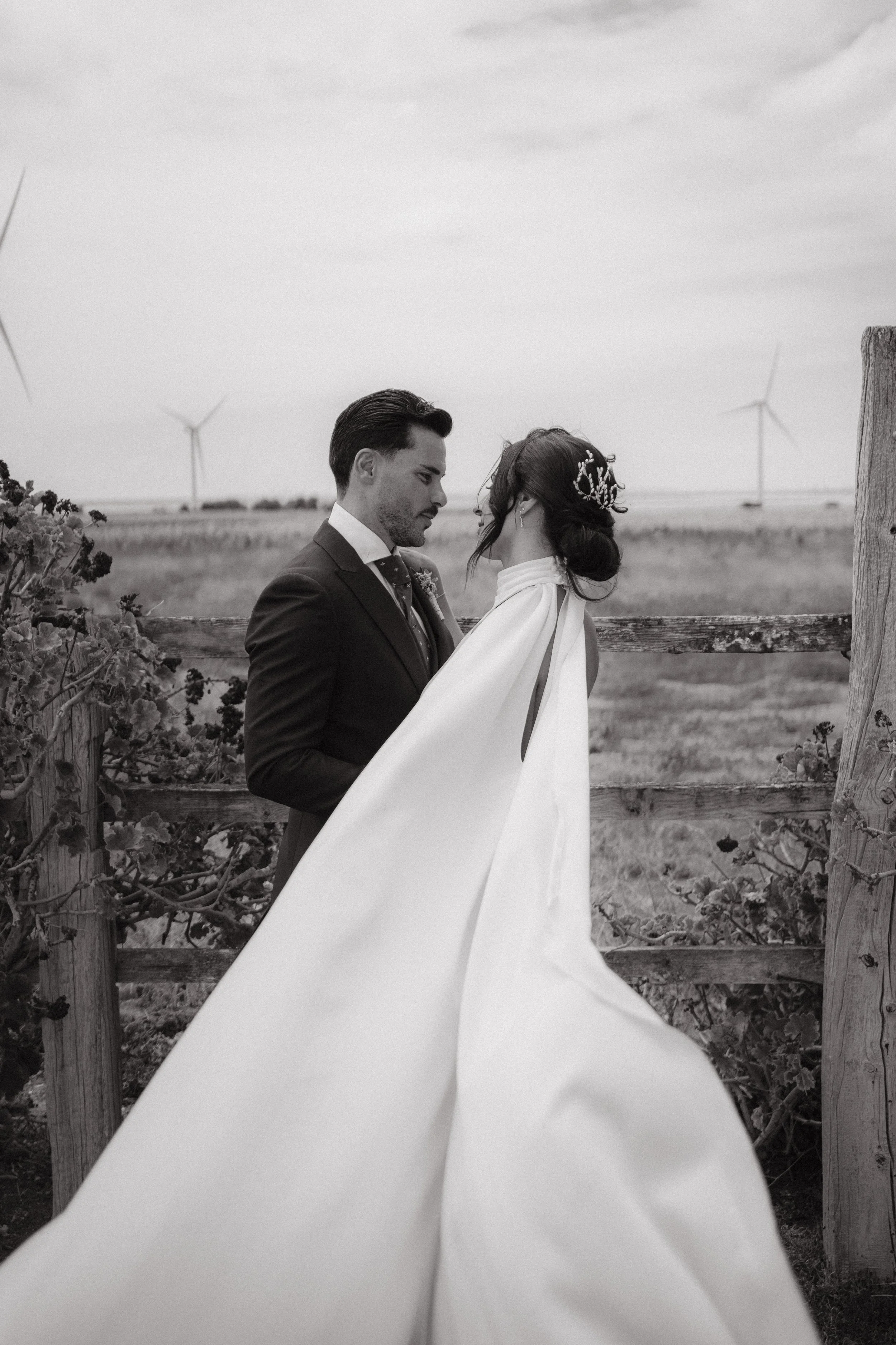 Pareja de recién casados frente a una cerca en campo abierto, con molinos de viento en el fondo.